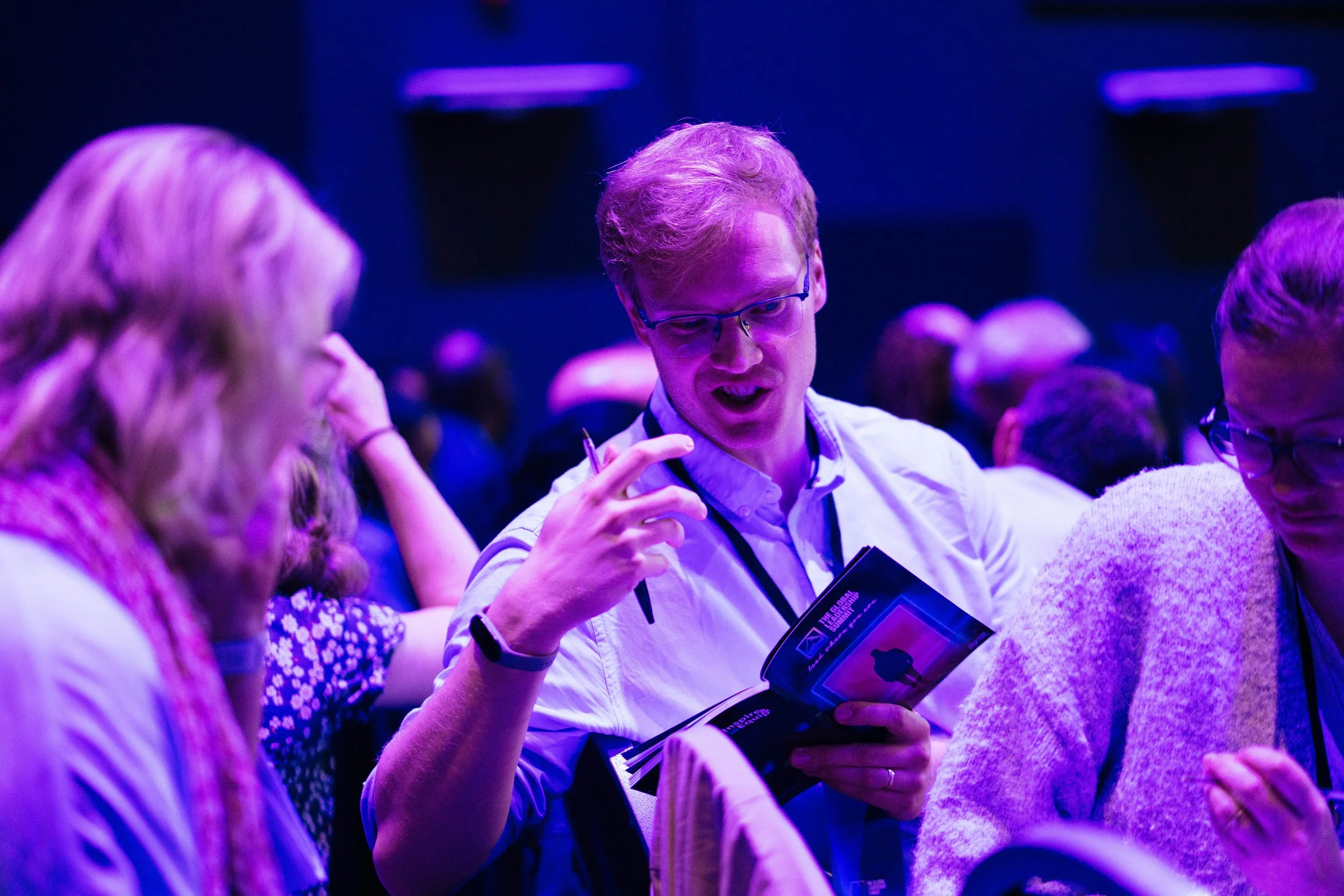 A group of people engaged in conversation at an indoor event with purple lighting. One man is holding a brochure and gesturing while talking.