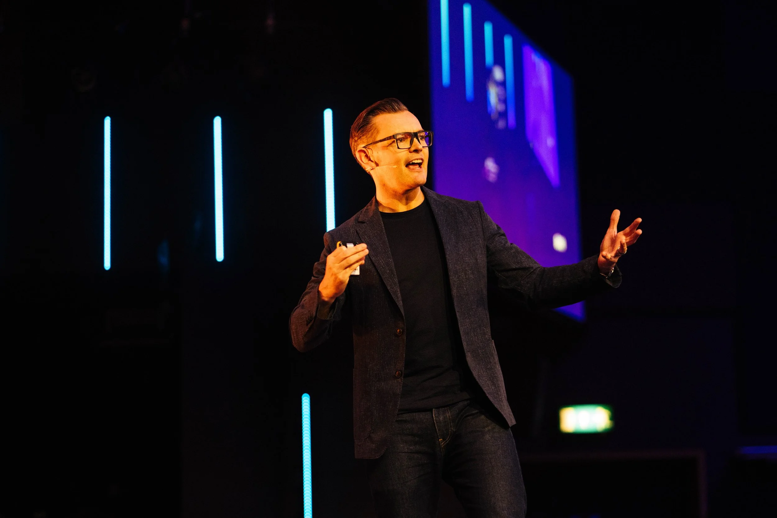 A man wearing glasses, a black shirt, and a dark blazer giving a presentation on a stage with blue neon vertical lights in the background.