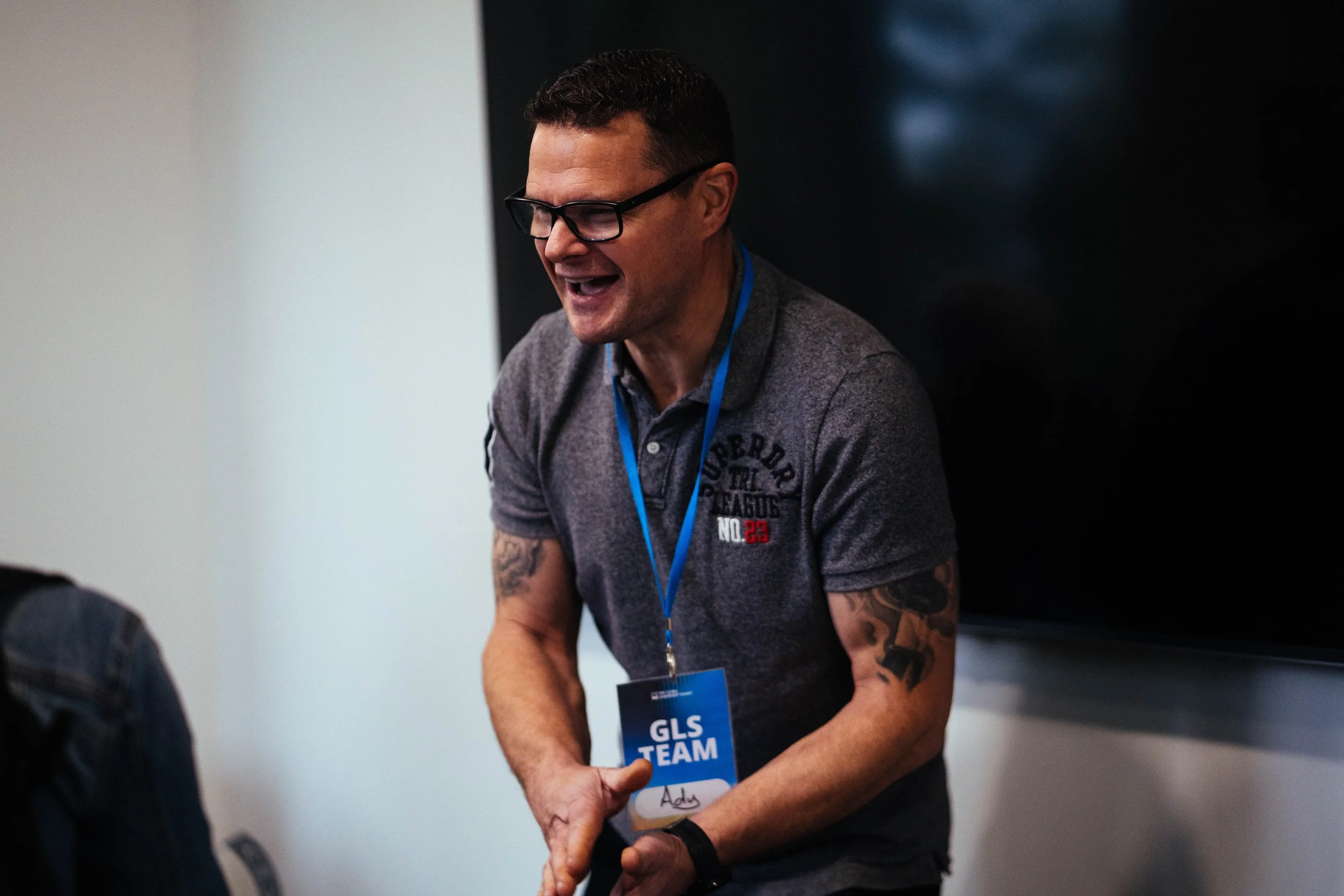 Man with glasses and tattoos wearing a gray shirt with a name tag that reads 'Adv' at a professional event, smiling and interacting with others.