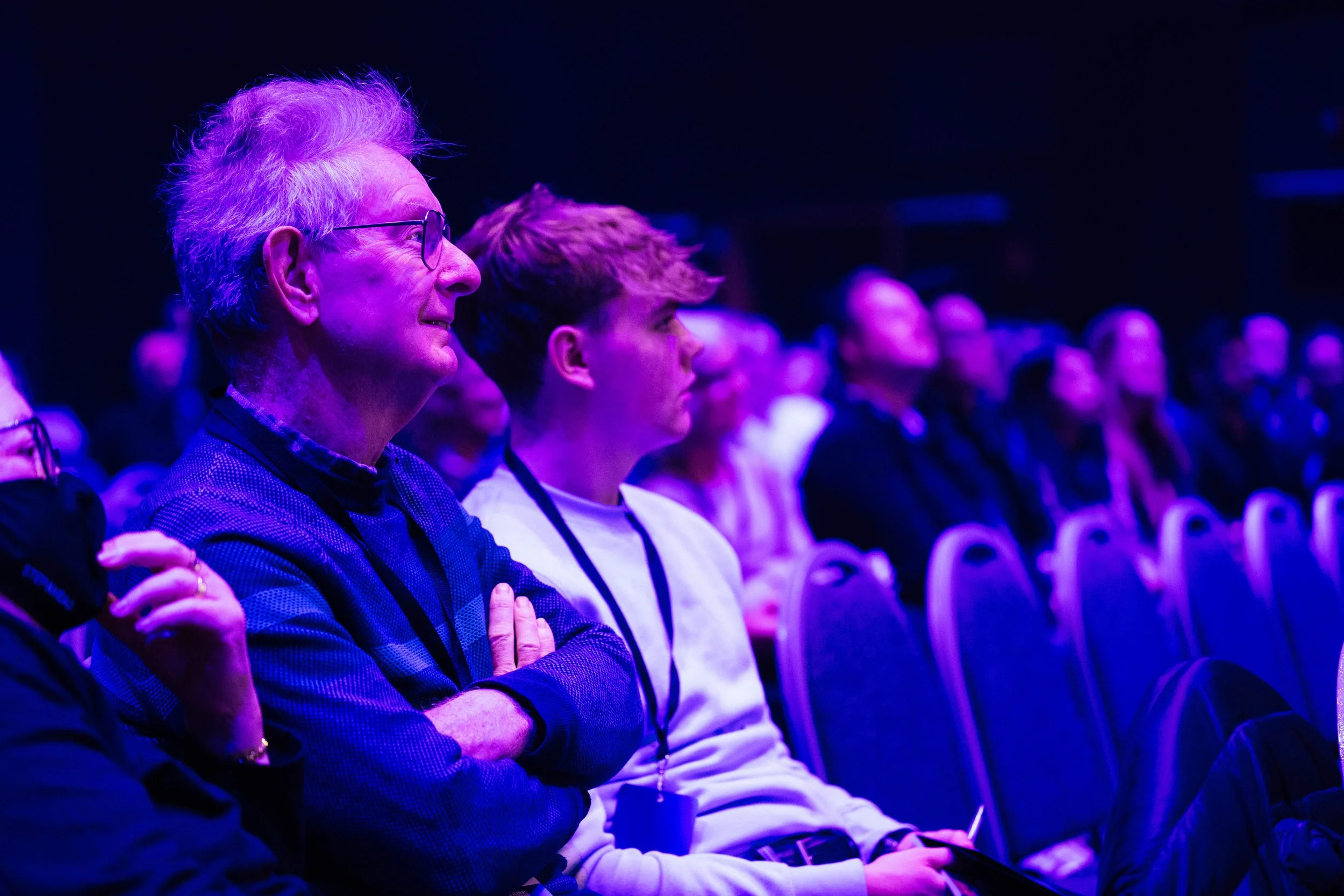 Audience members attentively listening at an event illuminated with purple and blue lighting.