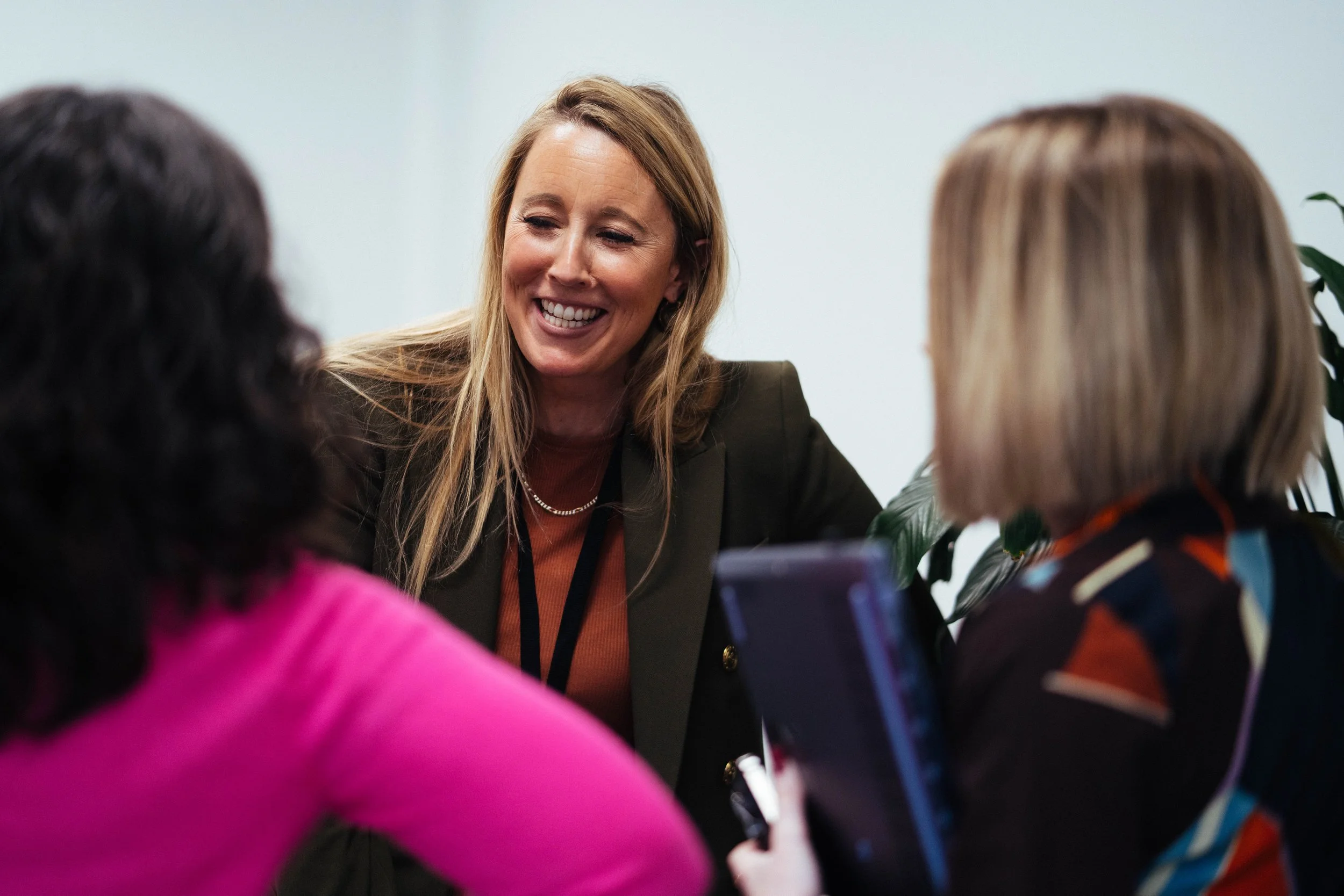 A woman with long blonde hair smiling and talking to two children, one wearing a pink jacket and the other a colorful jacket, in an indoor setting.