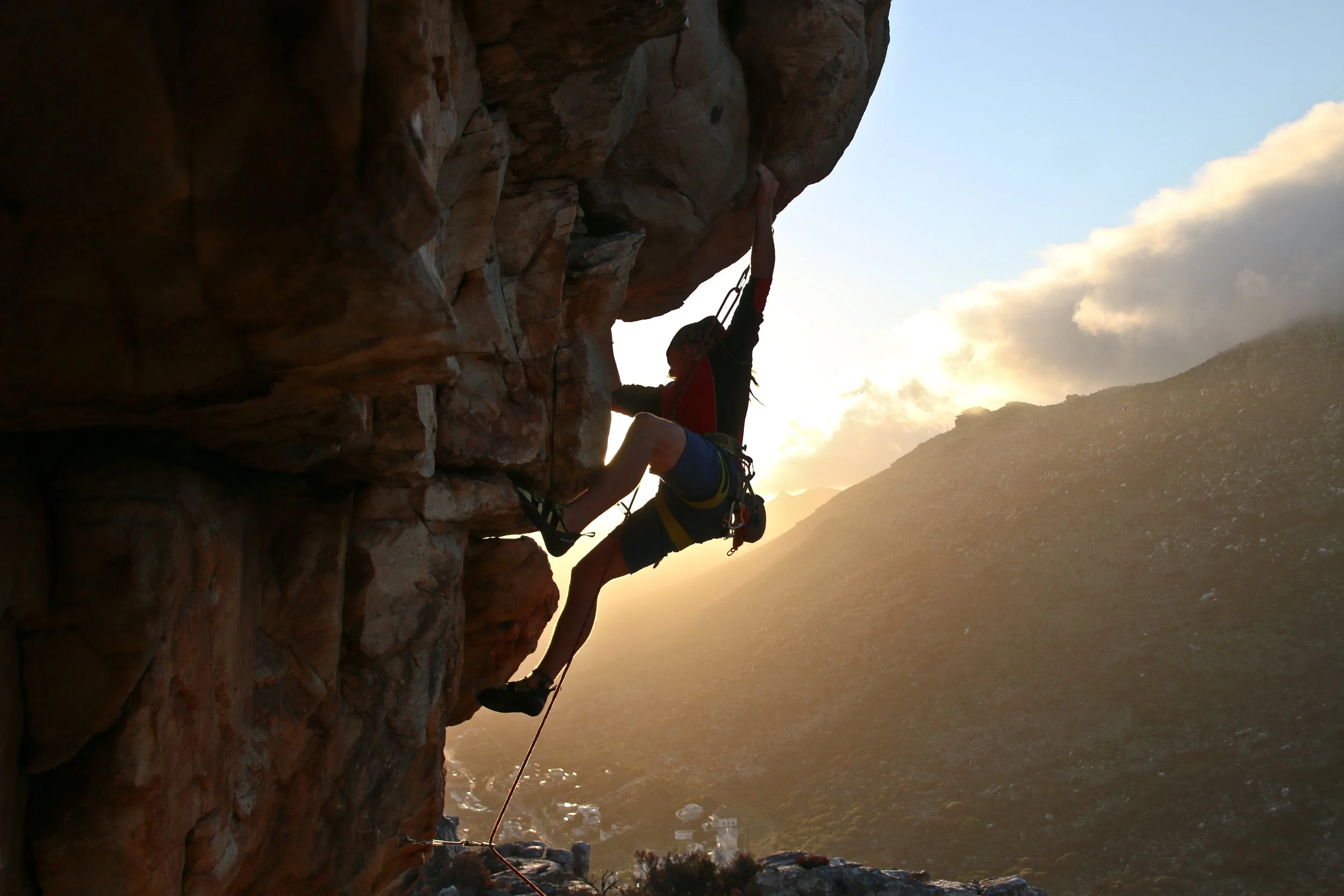 Person rock climbing on a steep cliff during sunset or sunrise with mountains and sky in the background.