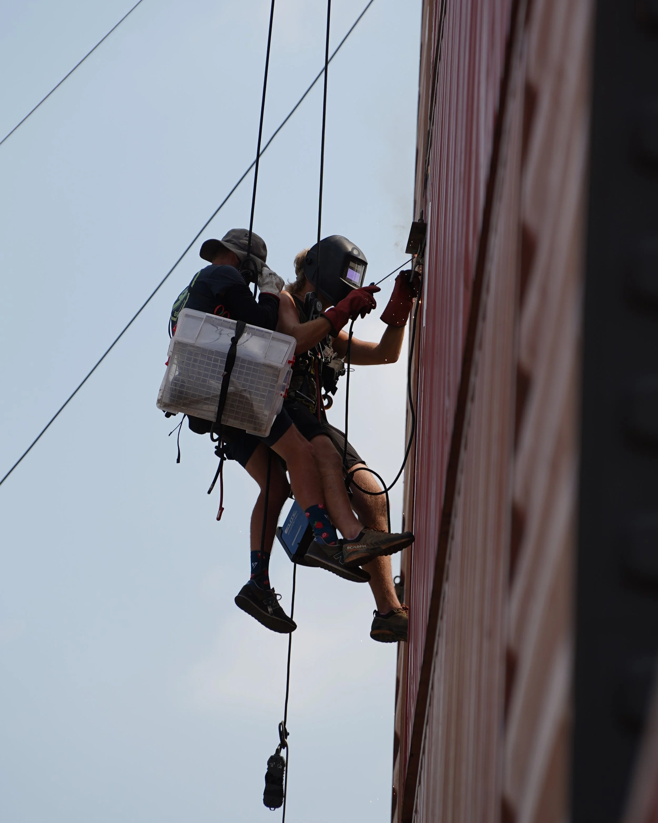 Two workers wearing safety gear, including helmets and harnesses, are installing or repairing a large red panel attached to a building exterior, with one worker assisting the other.
