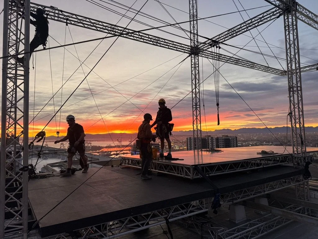 Construction workers setting up a stage on top of a high-rise building during sunset with a city skyline in the background.