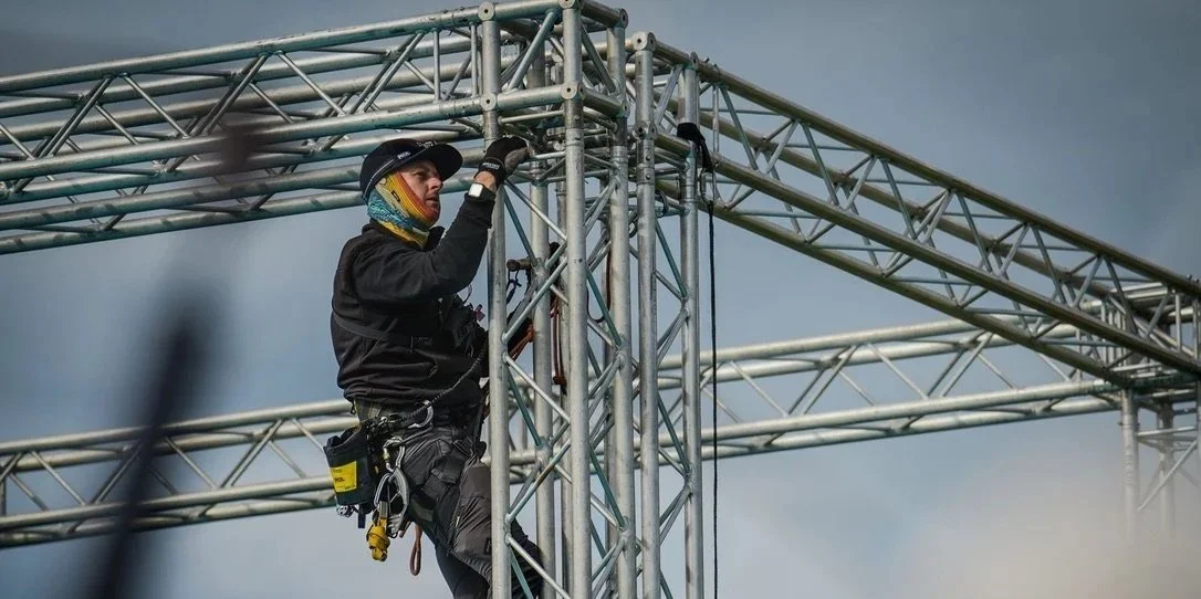 A worker in black clothing and safety gear is atop metal scaffolding, holding onto the structure as they work on assembling or maintaining the metal framework.