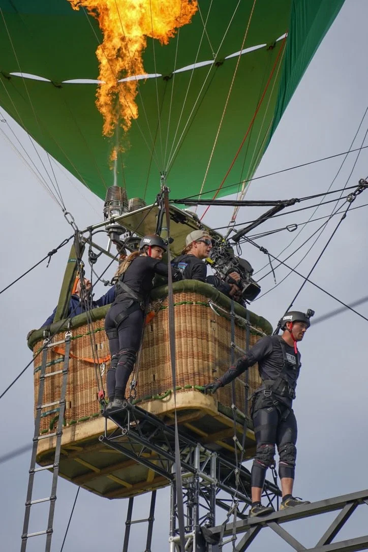 A hot air balloon with a green envelope and flame shooting up from the burner, with people inside the basket preparing for a flight, some standing outside the basket.