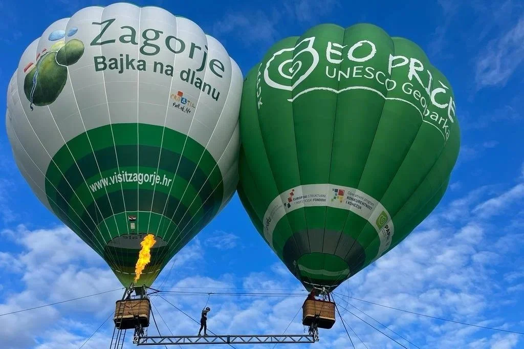 Two hot air balloons floating in the sky with clouds, one green and one multicolored, displaying logos and text in Croatian, with people in baskets below.