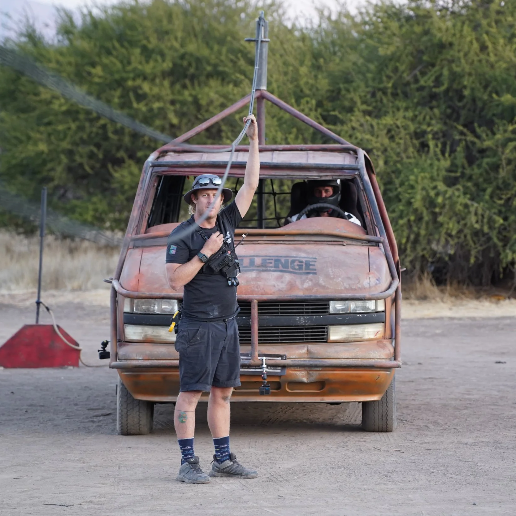 A man wearing a wide hat, sunglasses, a black t-shirt, and shorts stands in front of an old, weathered race car with a cage structure, holding a tool and a walkie-talkie. There is a person in a helmet sitting inside the car, and a red object with a handle is on the ground nearby. The background shows green trees and a dirt or gravel surface.