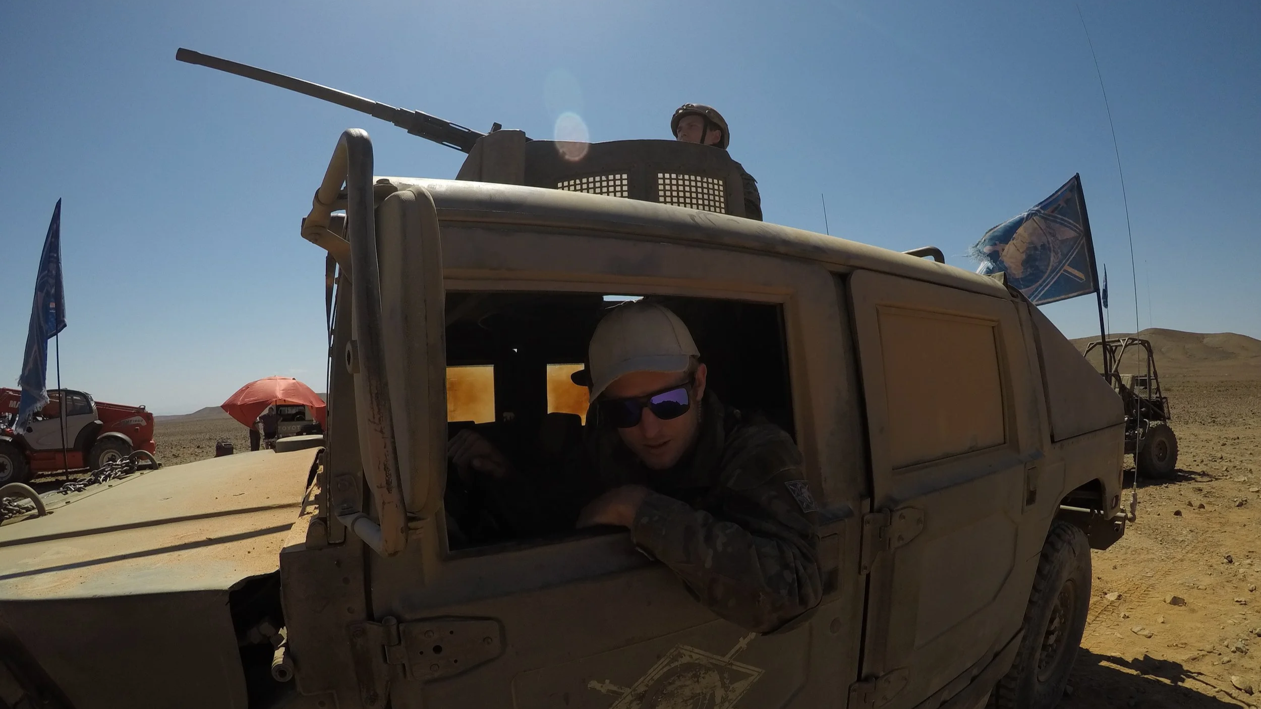 Soldiers inside a military vehicle in a desert with flags, another vehicle, and a person holding an umbrella in the background.