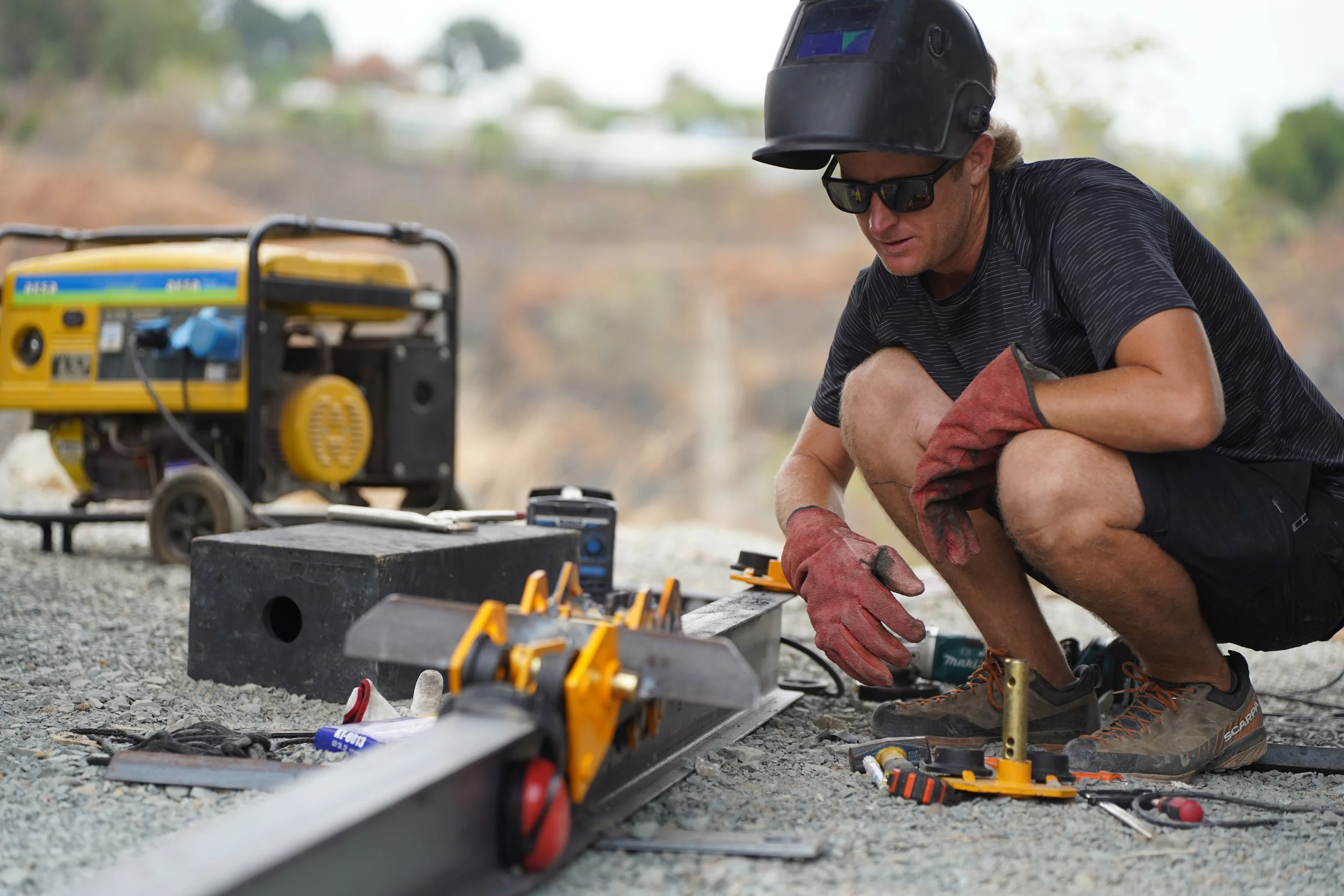 A construction worker kneeling on gravel, wearing a black helmet, sunglasses, and gloves, working with a level and tools outdoors with a generator in the background.