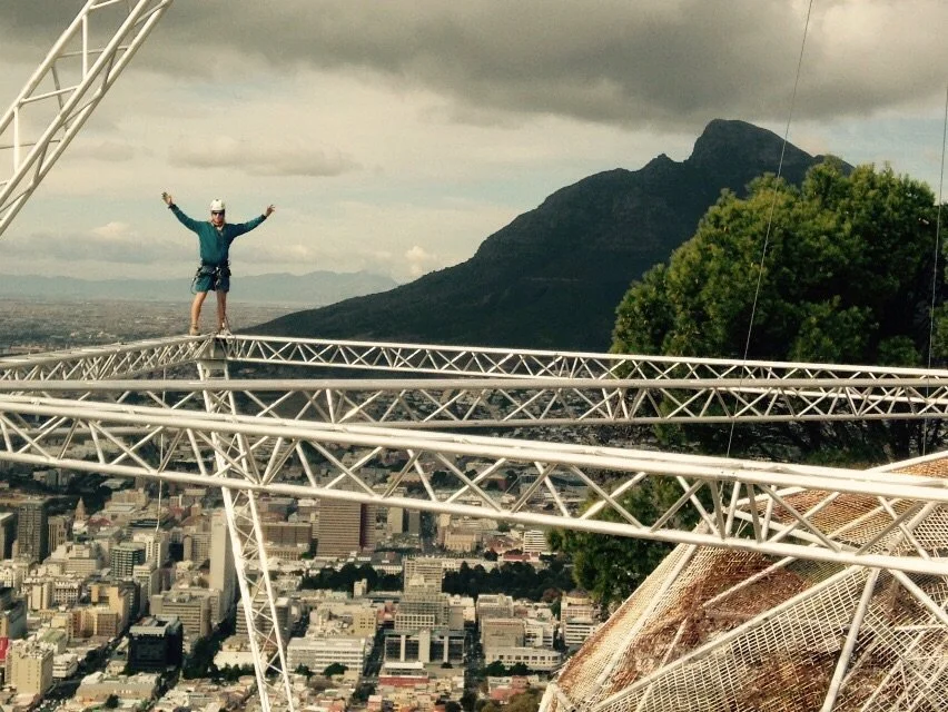 A person standing with arms raised on a narrow metal bridge high above a city with mountains in the background.