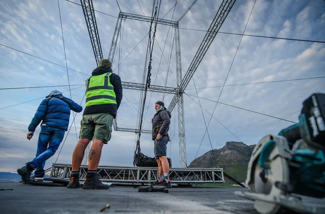 Three workers installing or maintaining a large outdoor electrical transmission tower during daytime with mountains and cloudy sky in background.