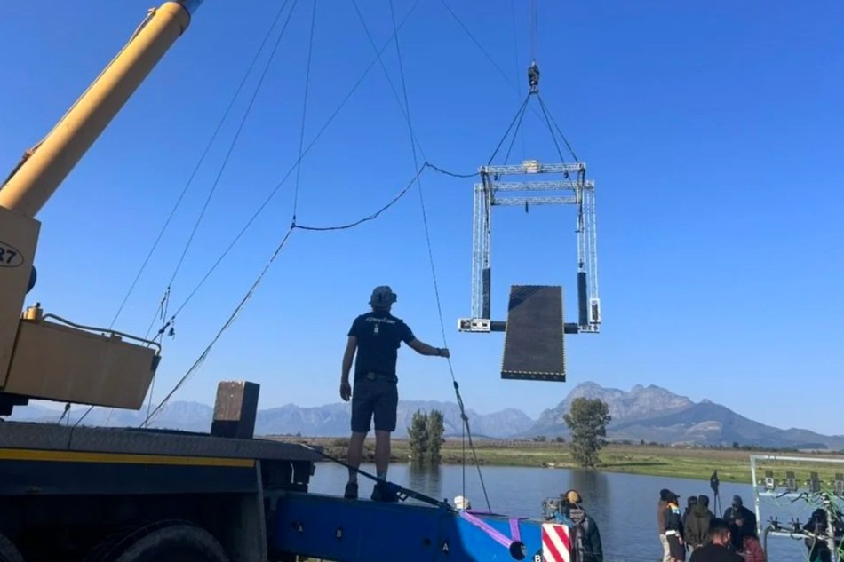 A person standing on a boat near a body of water, operating a crane that is lowering a large, empty metal frame with a platform at the bottom, with mountains in the background and a clear blue sky.