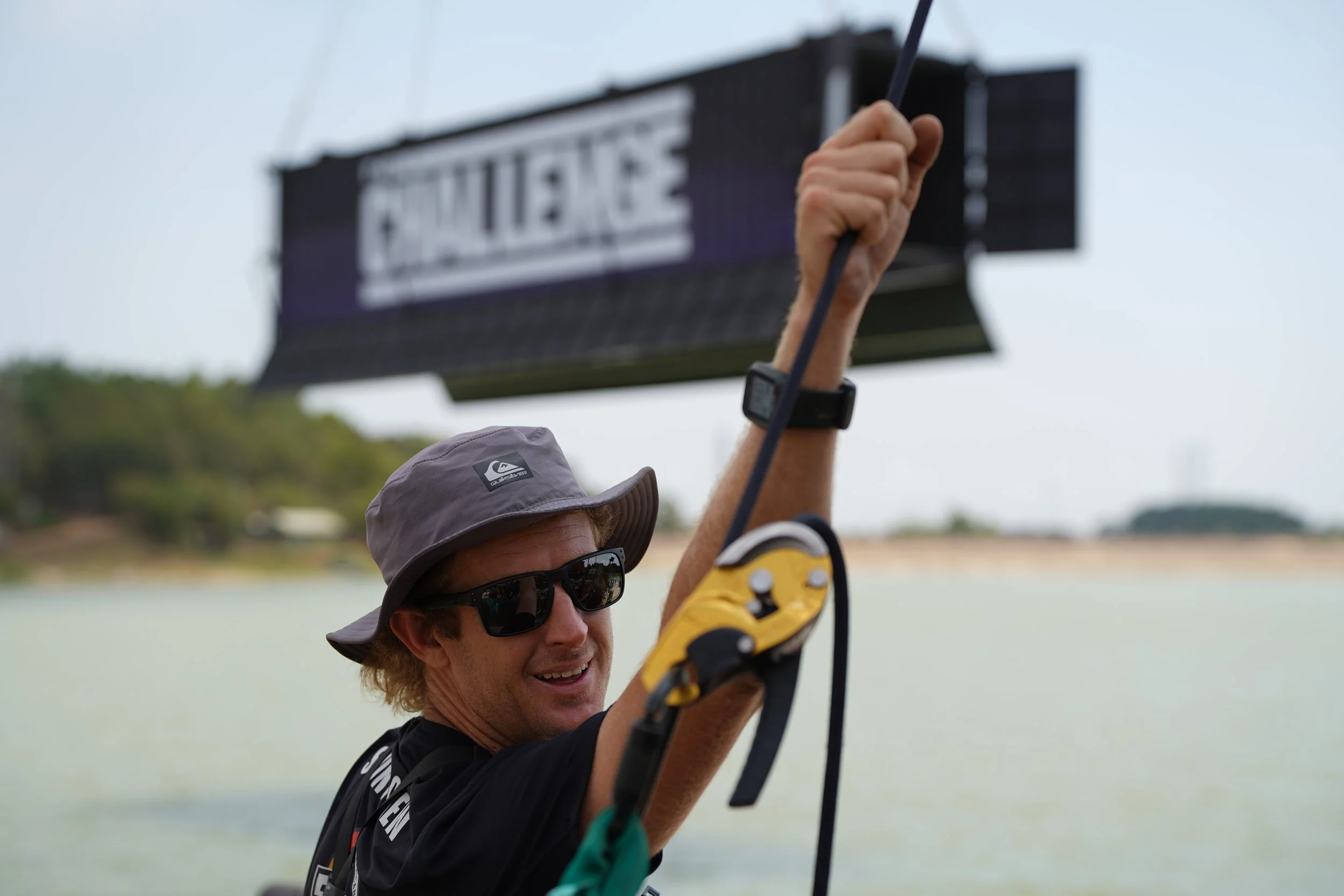 A person wearing sunglasses, a gray sun hat, and a black shirt, holding a fishing rod, smiling, outdoors near a body of water with trees in the background, with a blurry sign in the background.