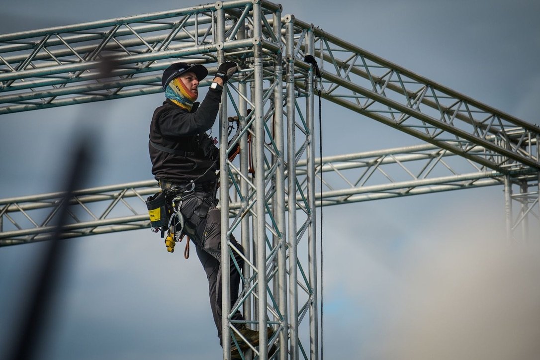 A worker wearing a safety harness and gloves is assembling or inspecting a metal scaffolding structure outdoors against a cloudy sky.