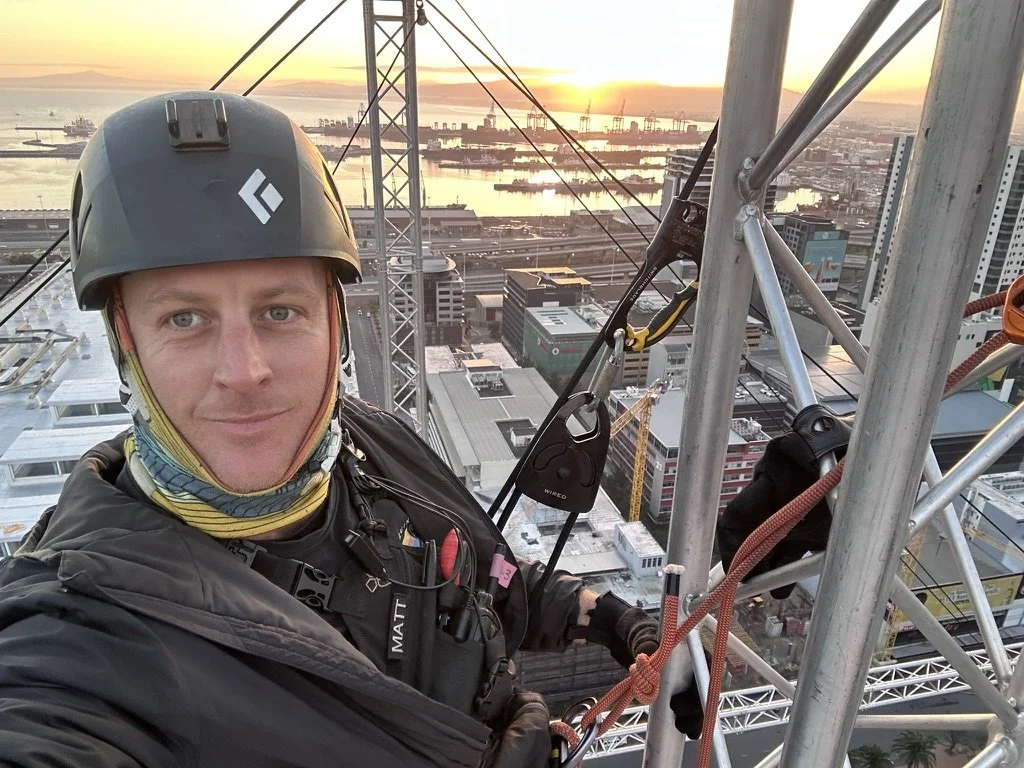 A man wearing climbing gear and a helmet takes a selfie from a high skyscraper at sunset, overlooking a city with water and cranes in the background.