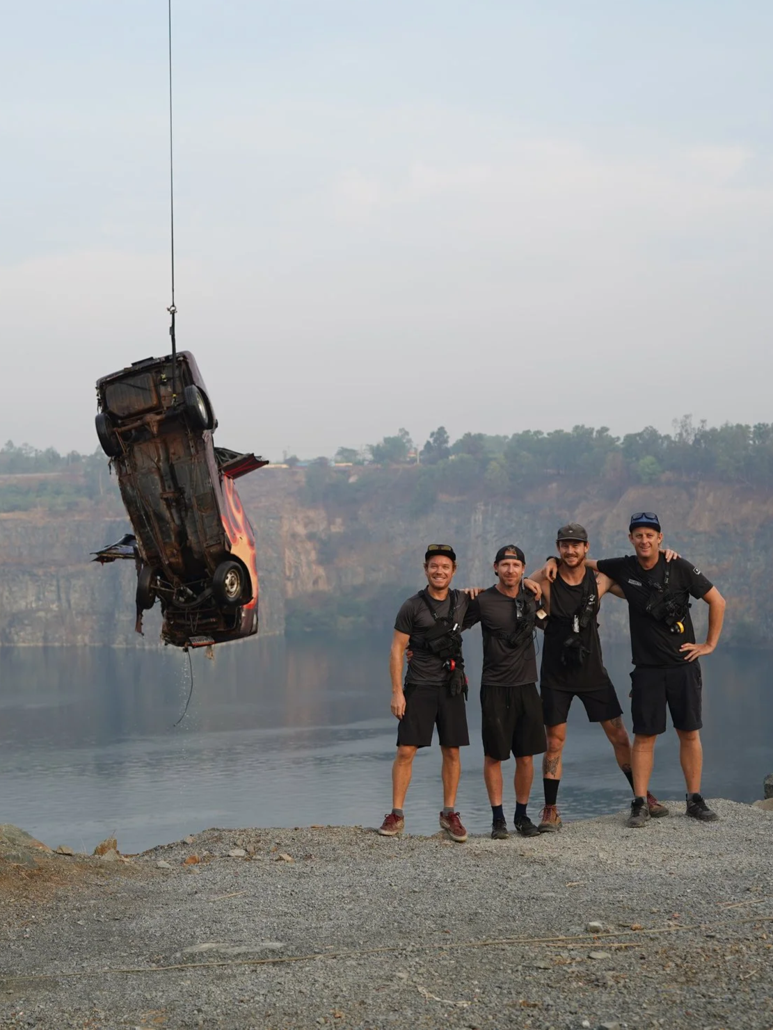 Four men stand side by side smiling with arms around each other on a rocky shore, while a damaged car is suspended in the air by a crane in the background.