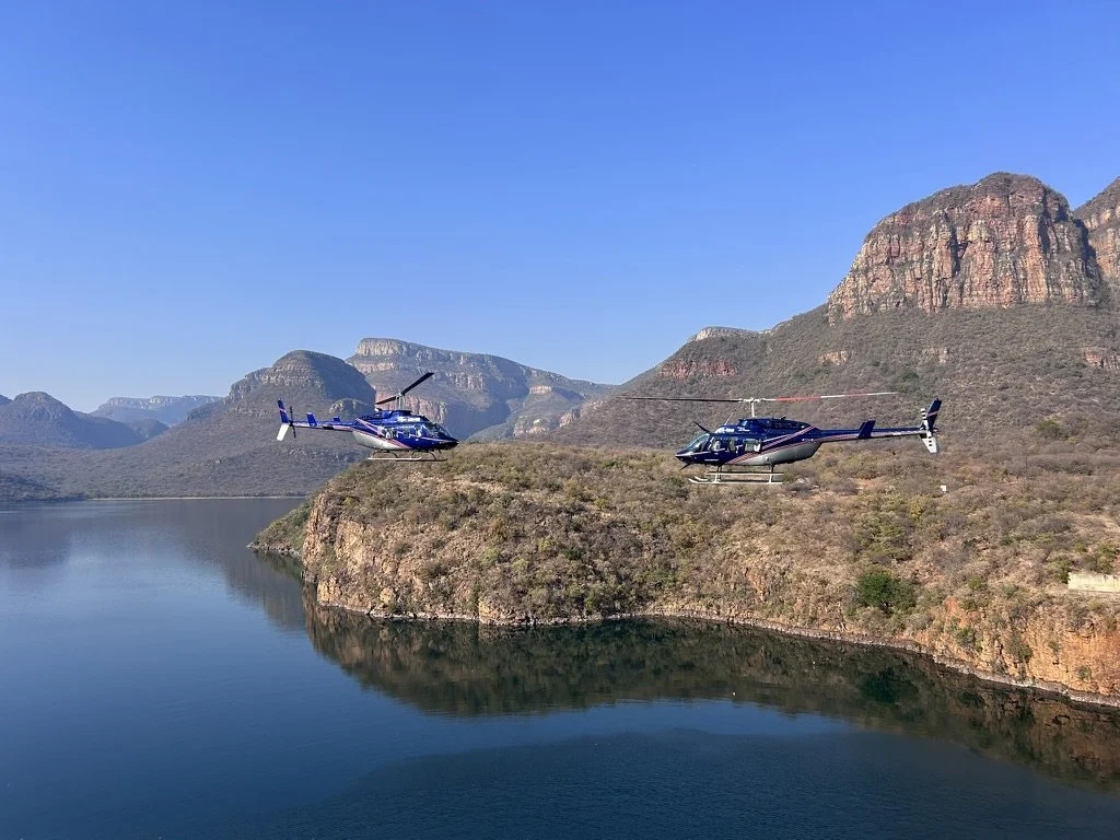 Two police helicopters flying over a large body of water with mountainous terrain in the background during daytime.