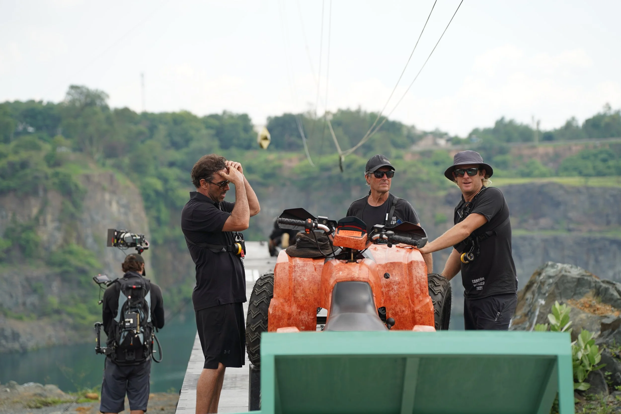 Three men are standing outdoors near a boat, some with sunglasses and hats. One man is touching his head, another is holding a tool, and the third is looking at a device on the boat. There are trees and hills in the background.