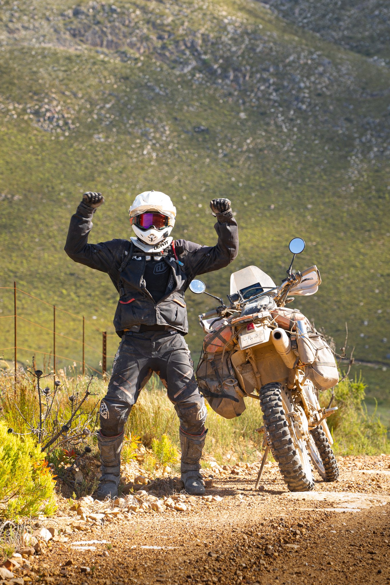 A motorcyclist in full gear, wearing a white helmet and black protective clothing, standing next to a dirt bike on a dirt road with a mountain background. The motorcyclist is raising both arms, flexing their muscles in a victorious pose.
