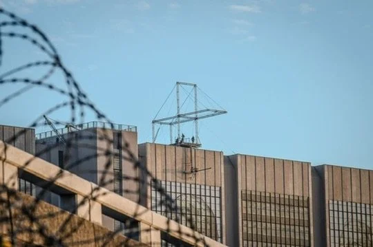 Cityscape with building, barbed wire fence, and transmission tower against a clear blue sky.