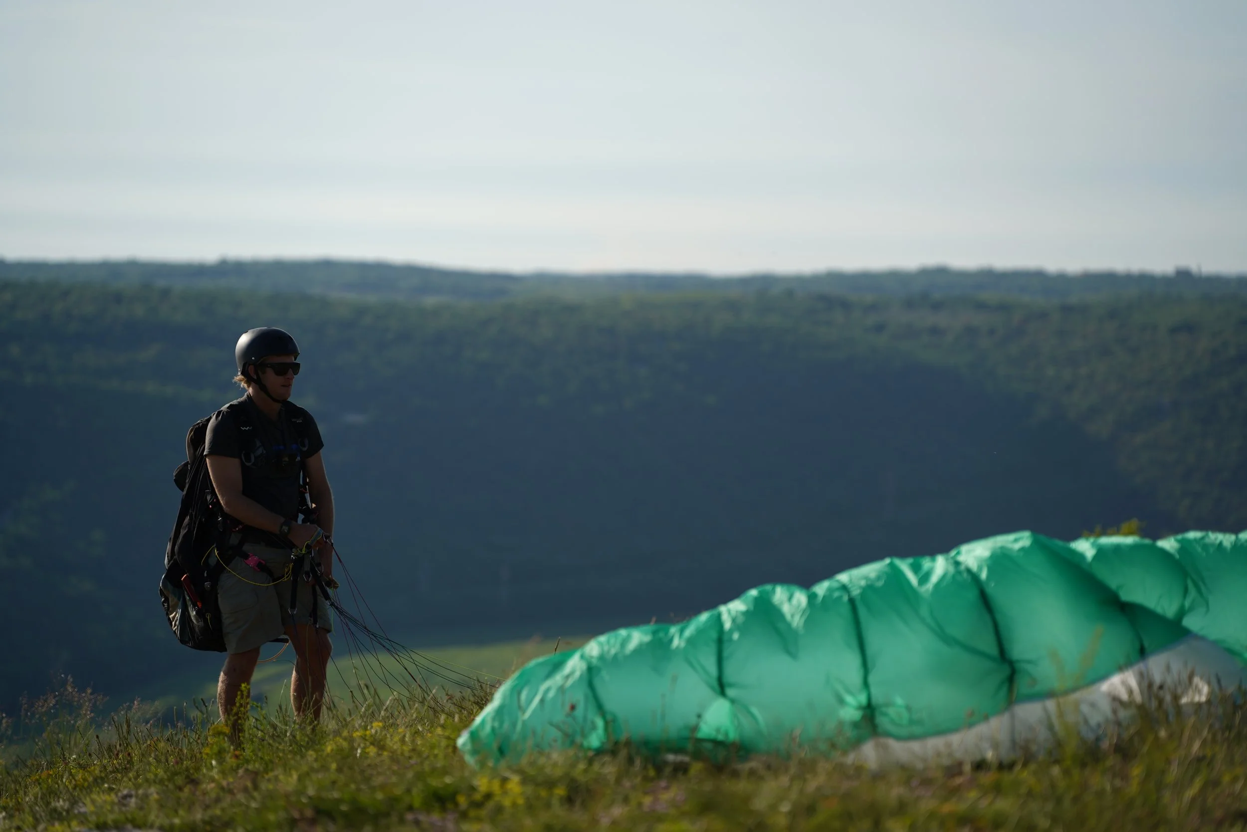 Man wearing helmet and sunglasses standing outdoors in front of a green and white parachute on grassy terrain with distant hills under a cloudy sky.