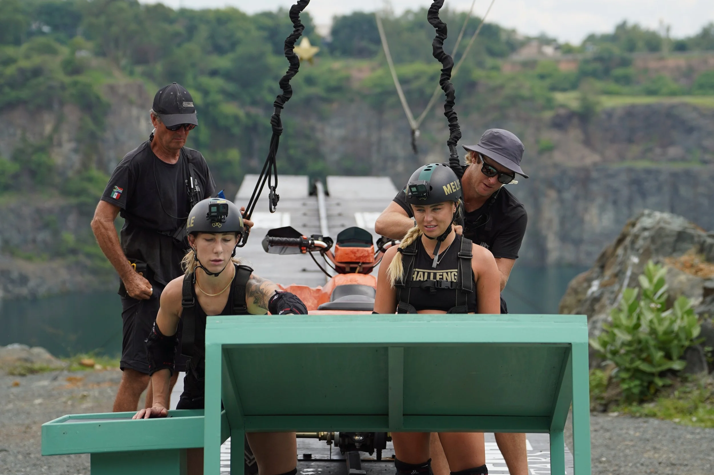 Two women with helmets and life jackets on a raft, with two men standing behind them, helping them with safety gear. The scene is set outdoors near water with trees and hills in the background.