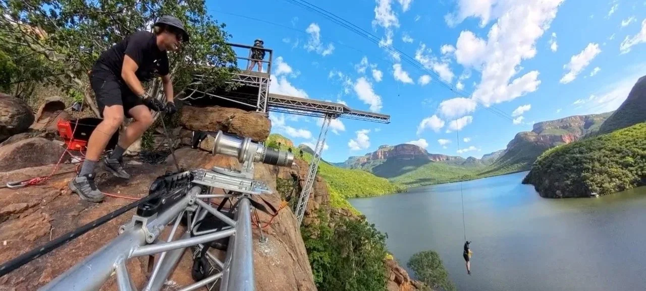 A person operating a camera on a tripod on a rocky ledge, with a person zip-lining across a river in a scenic mountainous area under a blue sky with clouds.