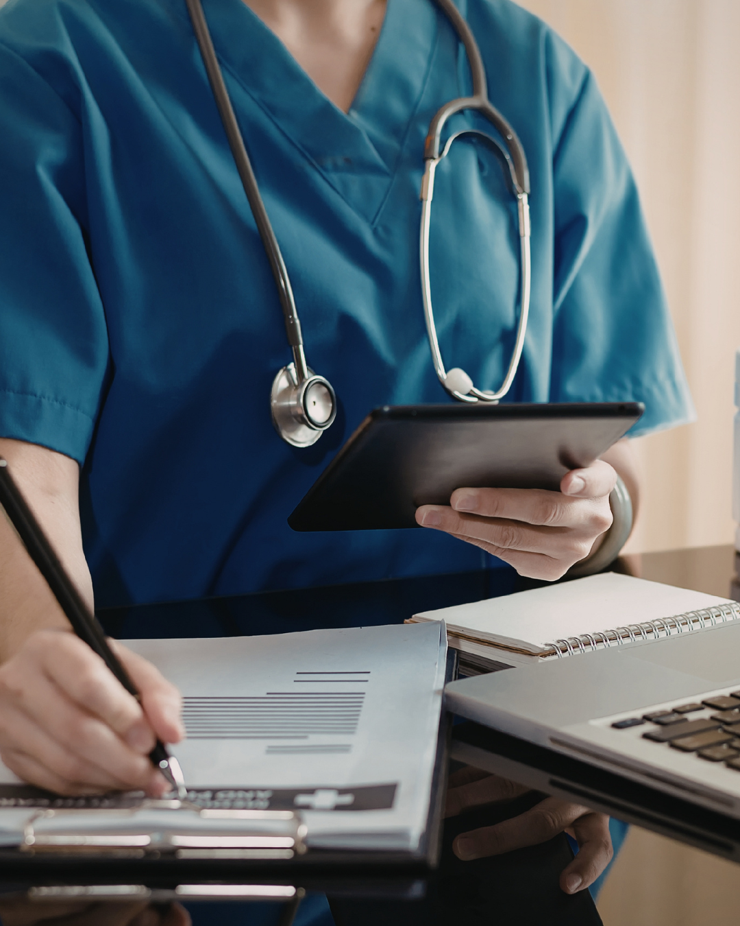 Healthcare professional in blue scrubs with a stethoscope using a tablet surrounded by medical documents and a laptop.