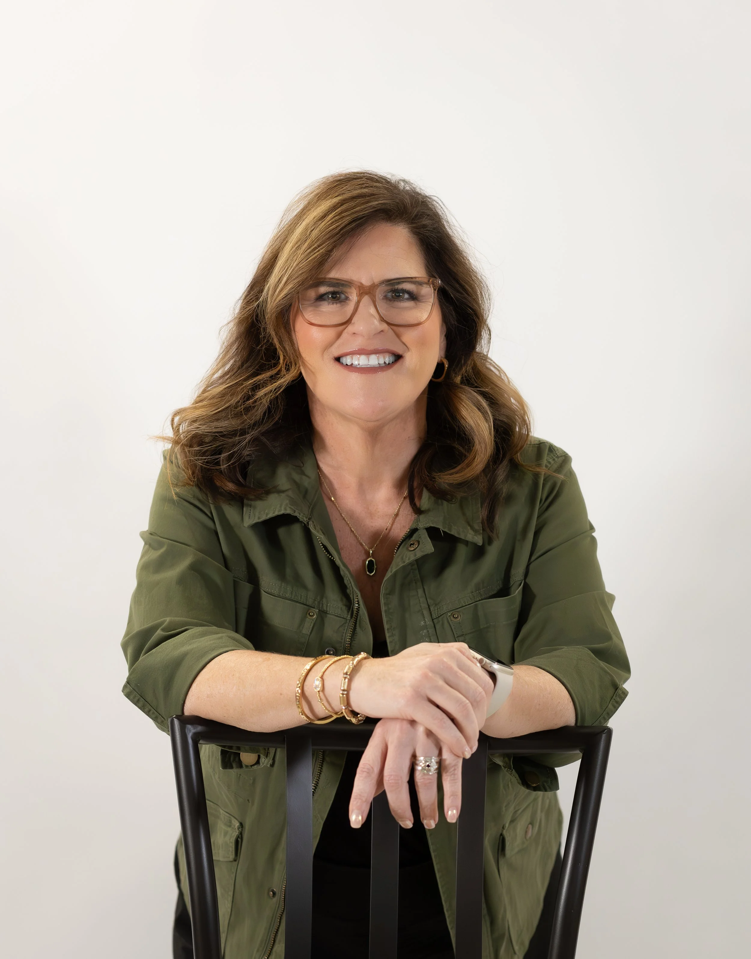Smiling woman with brown hair and glasses sitting at a black chair against a plain white background, wearing a green jacket and jewelry.