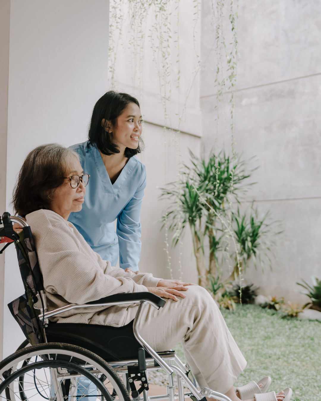 A young female nurse in blue scrubs talking to an elderly woman in a wheelchair, outdoors near a wall with greenery.