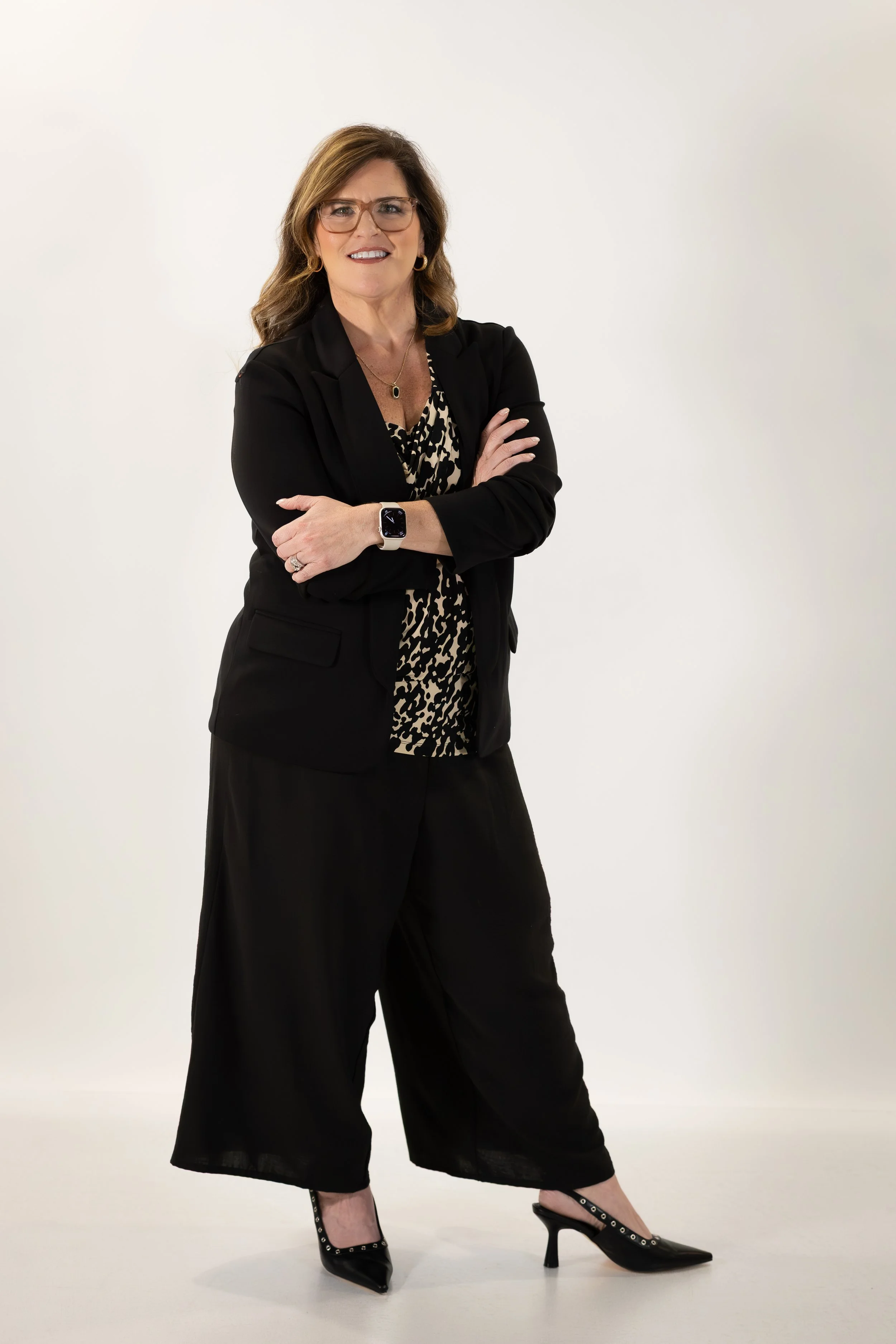 Woman in black blazer and wide-leg pants posing with crossed arms, wearing glasses, a leopard print blouse, and high heels with ankle strap details against a white background.