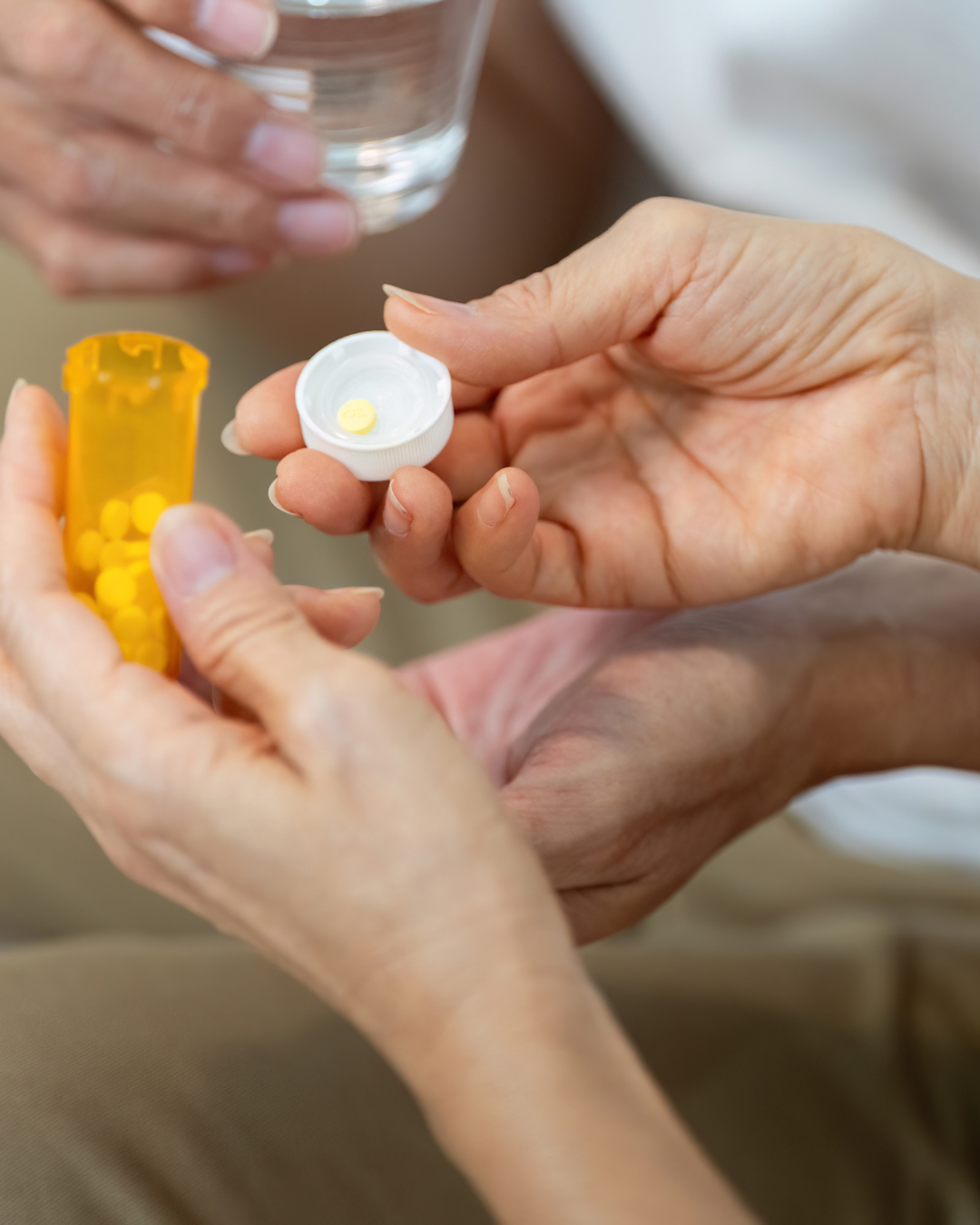 Close-up of a person's hand holding an open pill bottle with yellow pills and another hand holding a white pill organizer with yellow pills, with a glass of water in the background.