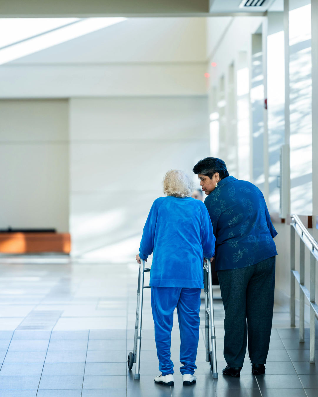 An elderly woman with white hair using a walker, receiving assistance from a younger woman in a hospital or care facility.