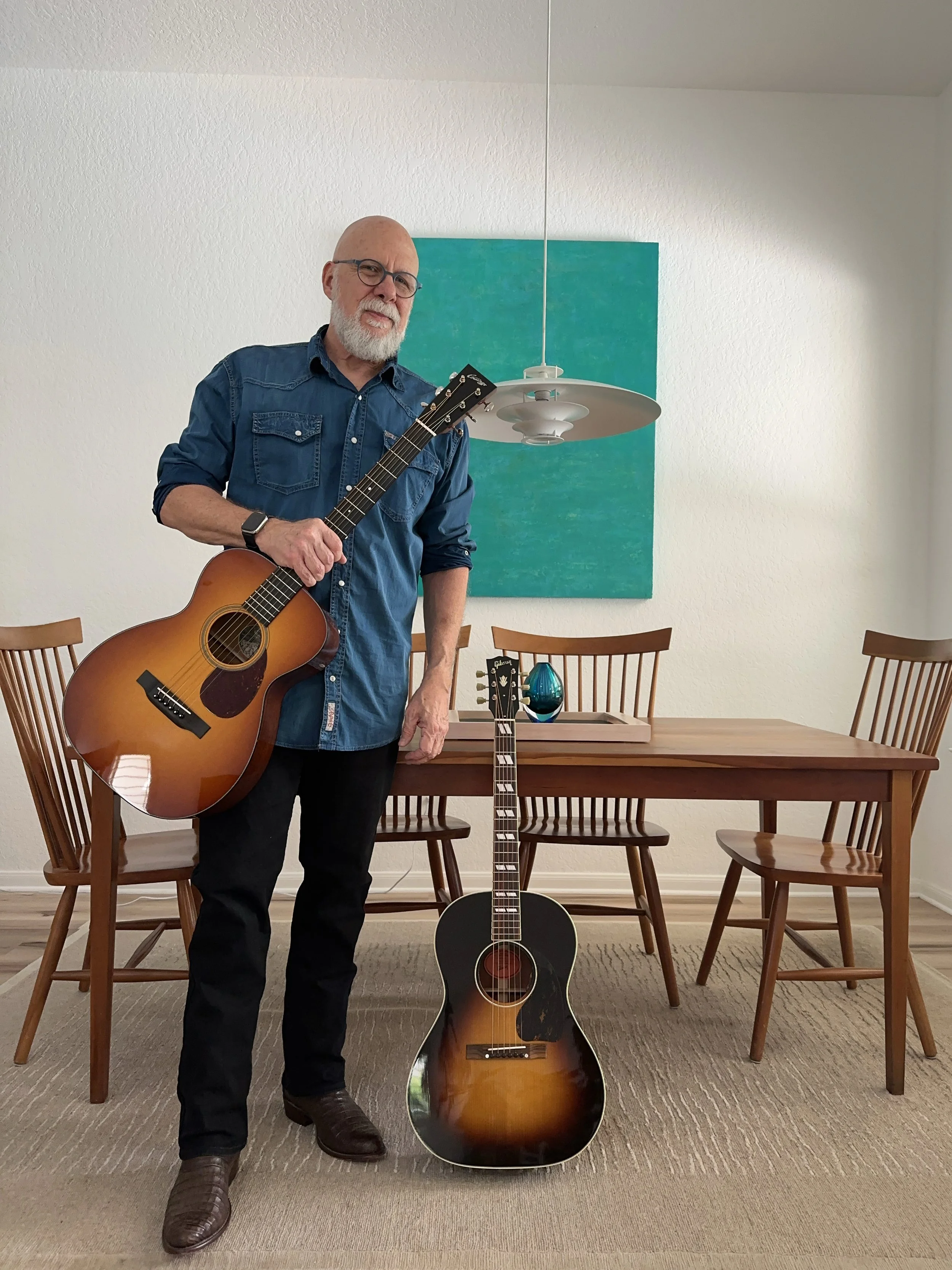 A man with glasses and a beard, wearing a denim shirt and black pants, holding an acoustic guitar, standing in a dining room with wooden chairs, a wooden table, and a teal wall hanging, and a black guitar on the floor in front of him.