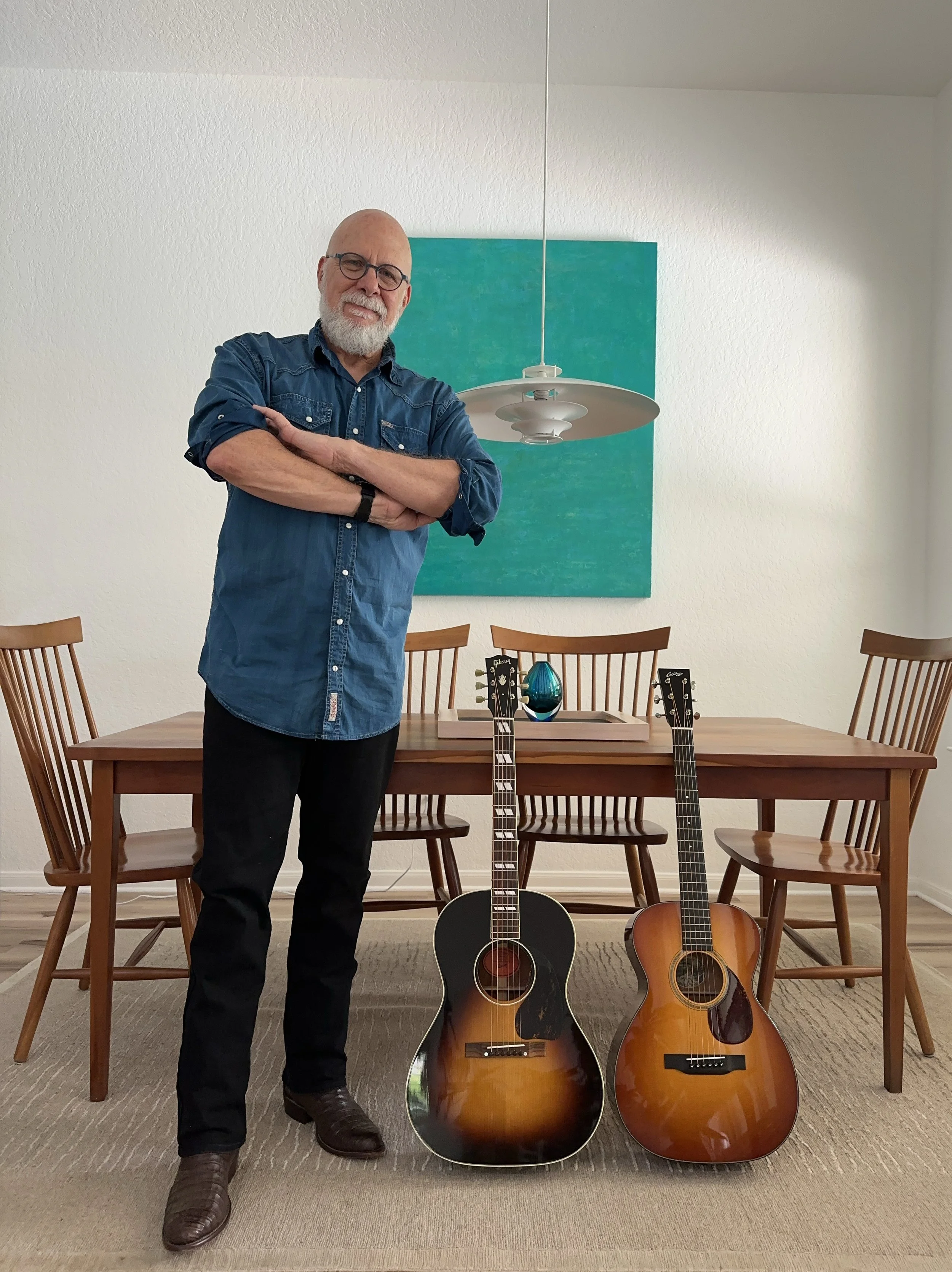 Older man with glasses and a beard standing with crossed arms in a dining room with three guitars in front of a wooden dining table, two are acoustic guitars and one appears to be a classical guitar. The man is smiling and wearing a blue shirt and dark pants. The background features a green painting and a white wall with a hanging light fixture.