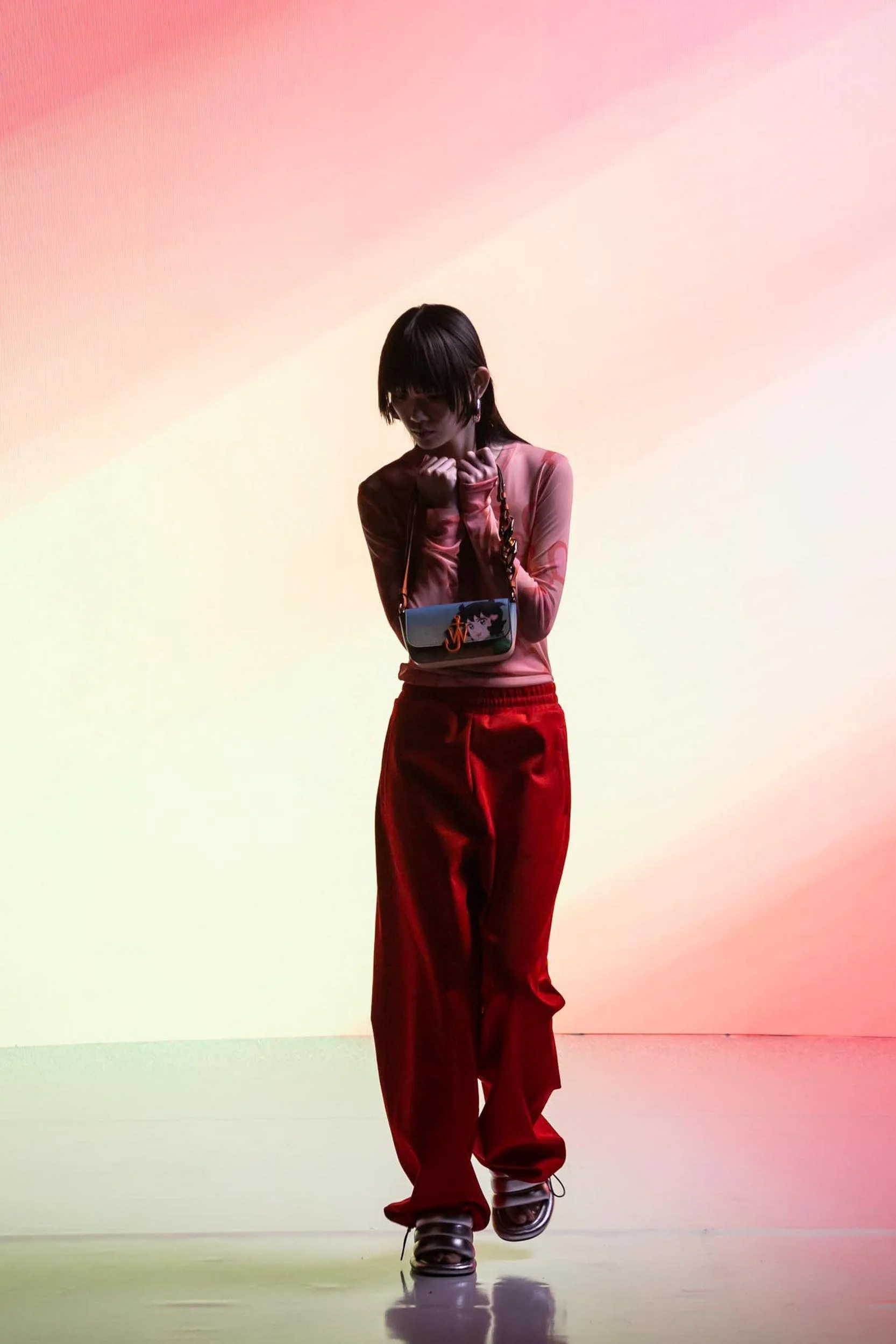 Female model posing with a hand bag with anime character on wearing bright red trousers and pink shirt with a reflective floor and abstract pinkish background shown on a large LED screen fashion shoot