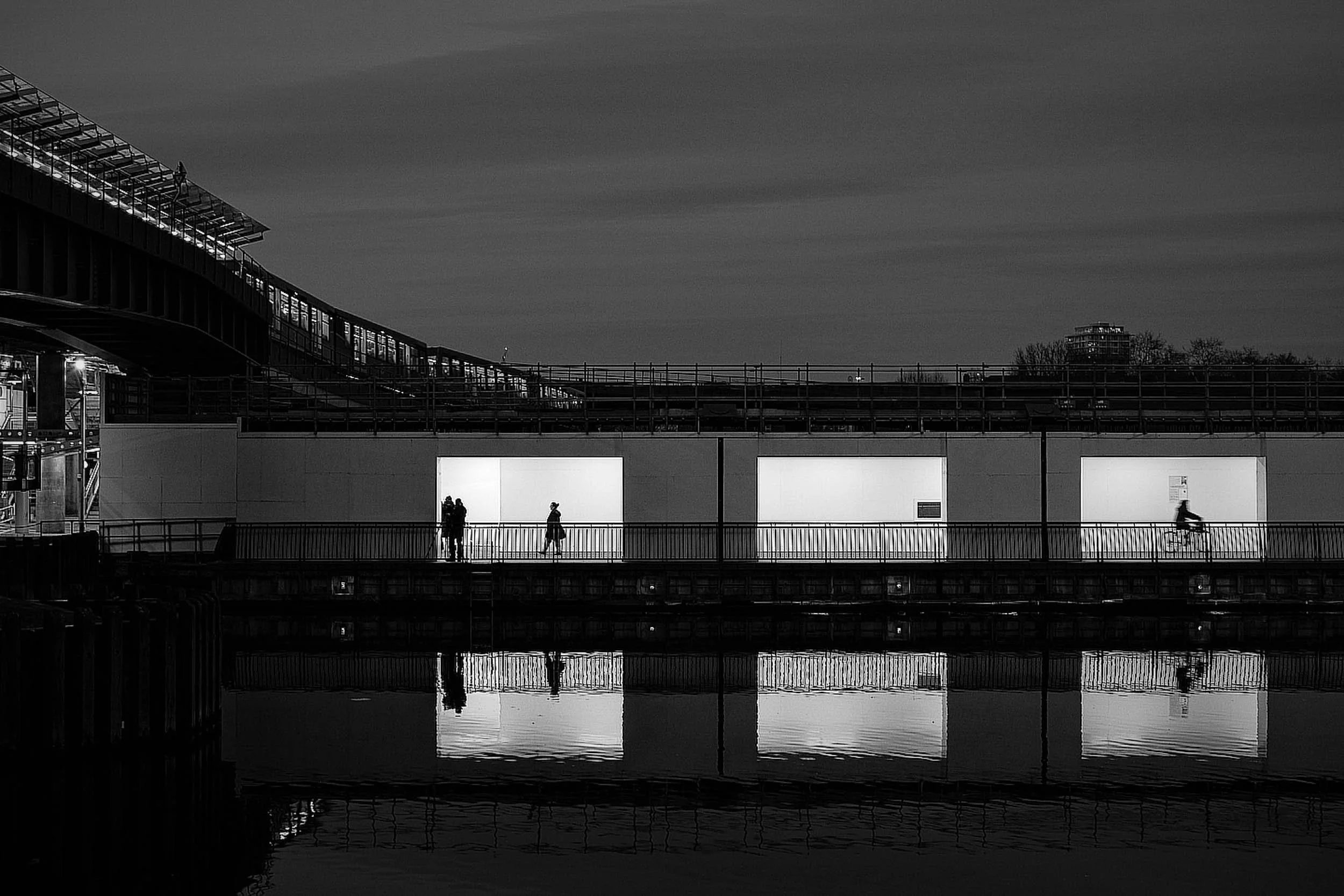 Black and white night scene looking over water to a neon lit walkway with silhouettes passing by in Canary Wharf London