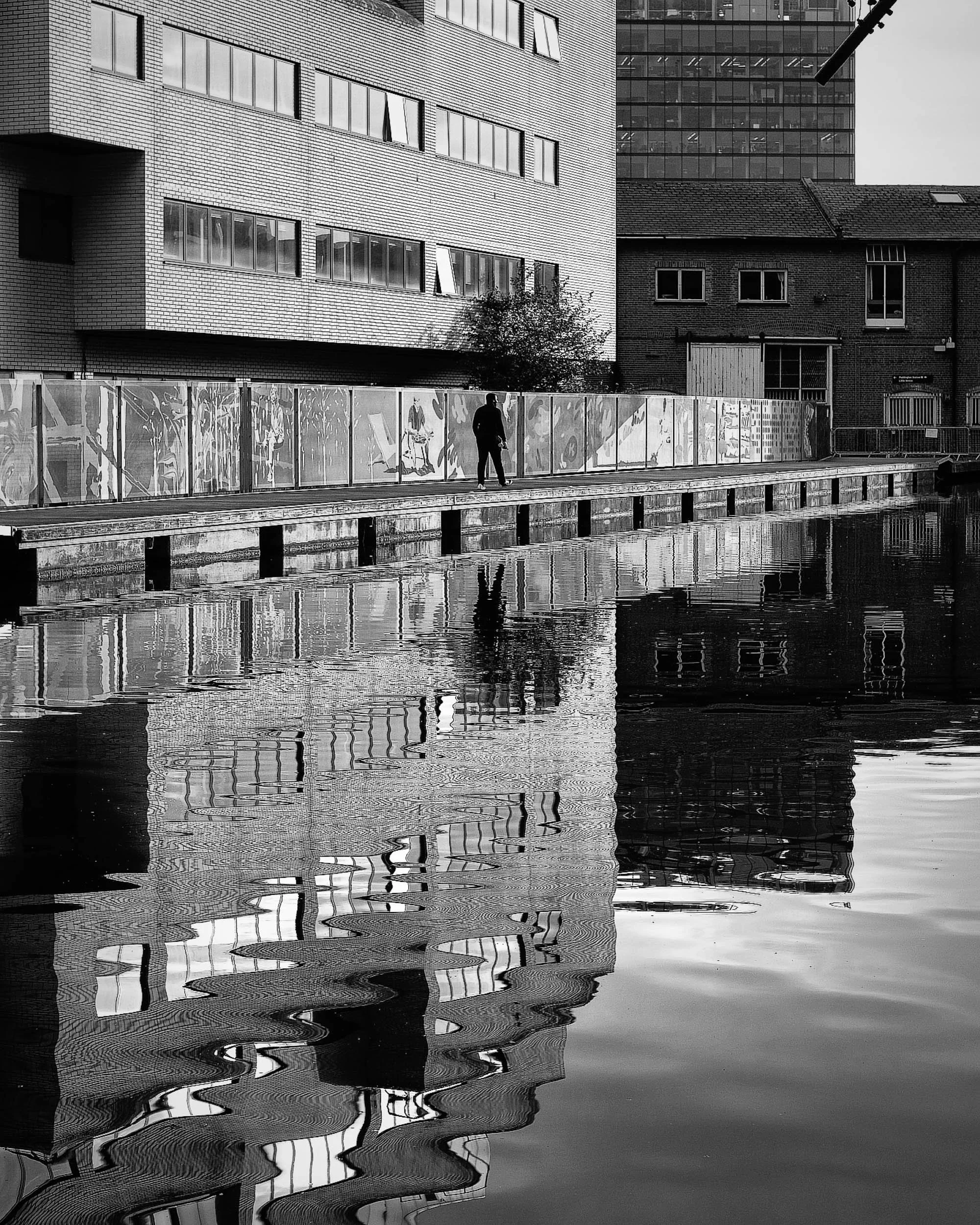 Black and white water reflections distort reality with ripples as a man walks by the canal in London