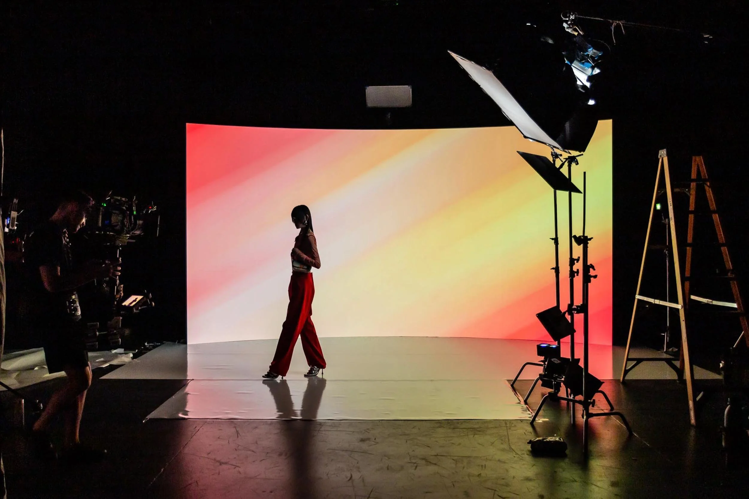 A person standing in a photo studio with a colorful gradient background on an LED Wall used for Virtual Production. Studio lights and equipment are visible, and the person is wearing red pants and a dark top.