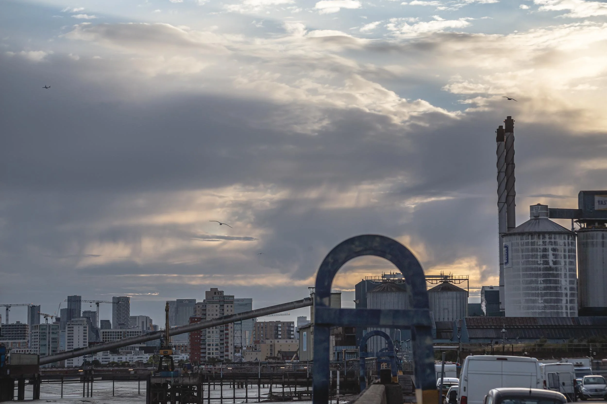 City skyline with tall buildings, cranes, and a cloudy sky, viewed from industrial estate on the side of the river in London with a view of Tate & Lyle Sugar Factory.