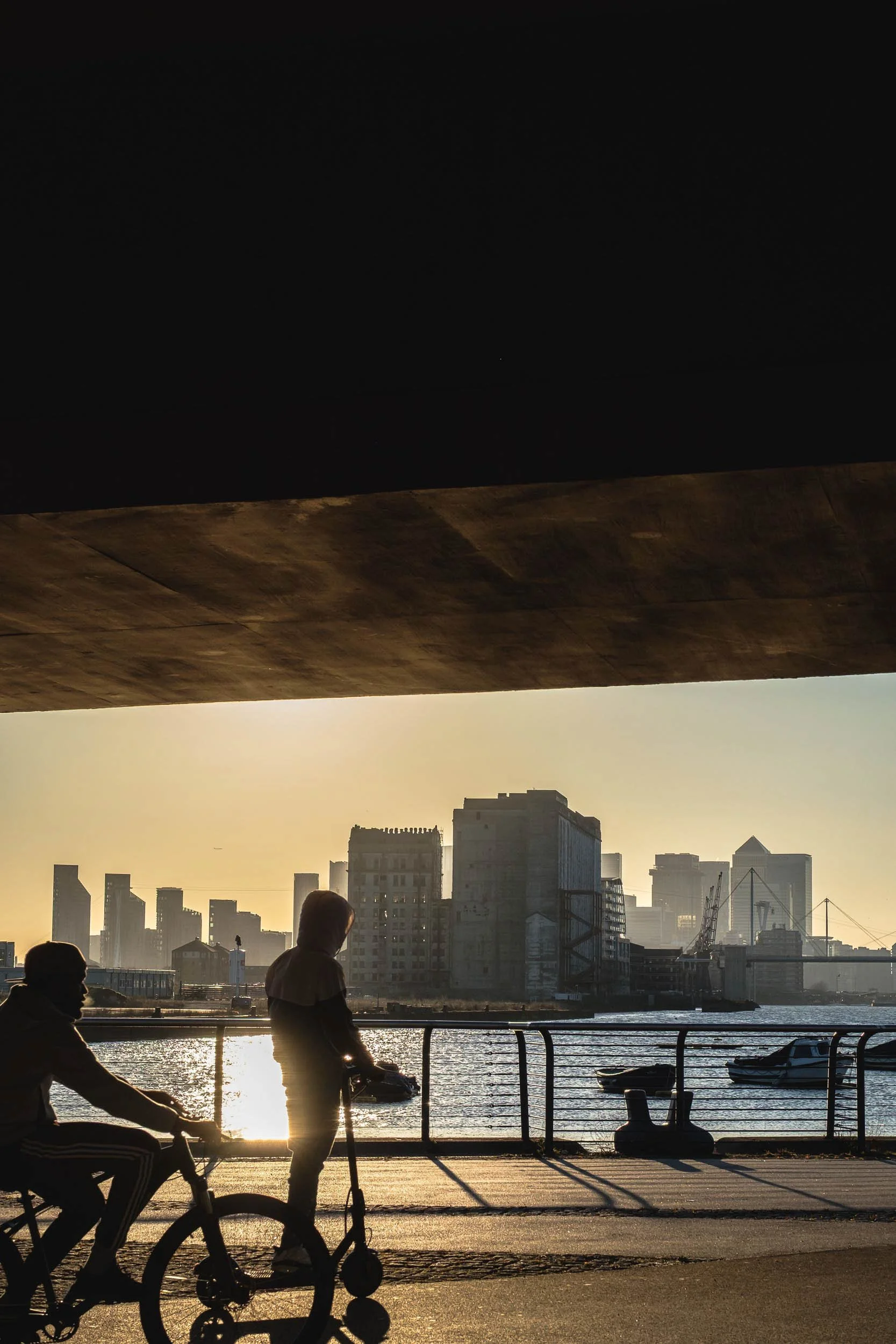 Golden hour in east London Royal Docks overlooking new building developments and Canary Wharf as silhouettes of people on bikes and escooters pass by the golden reflections
