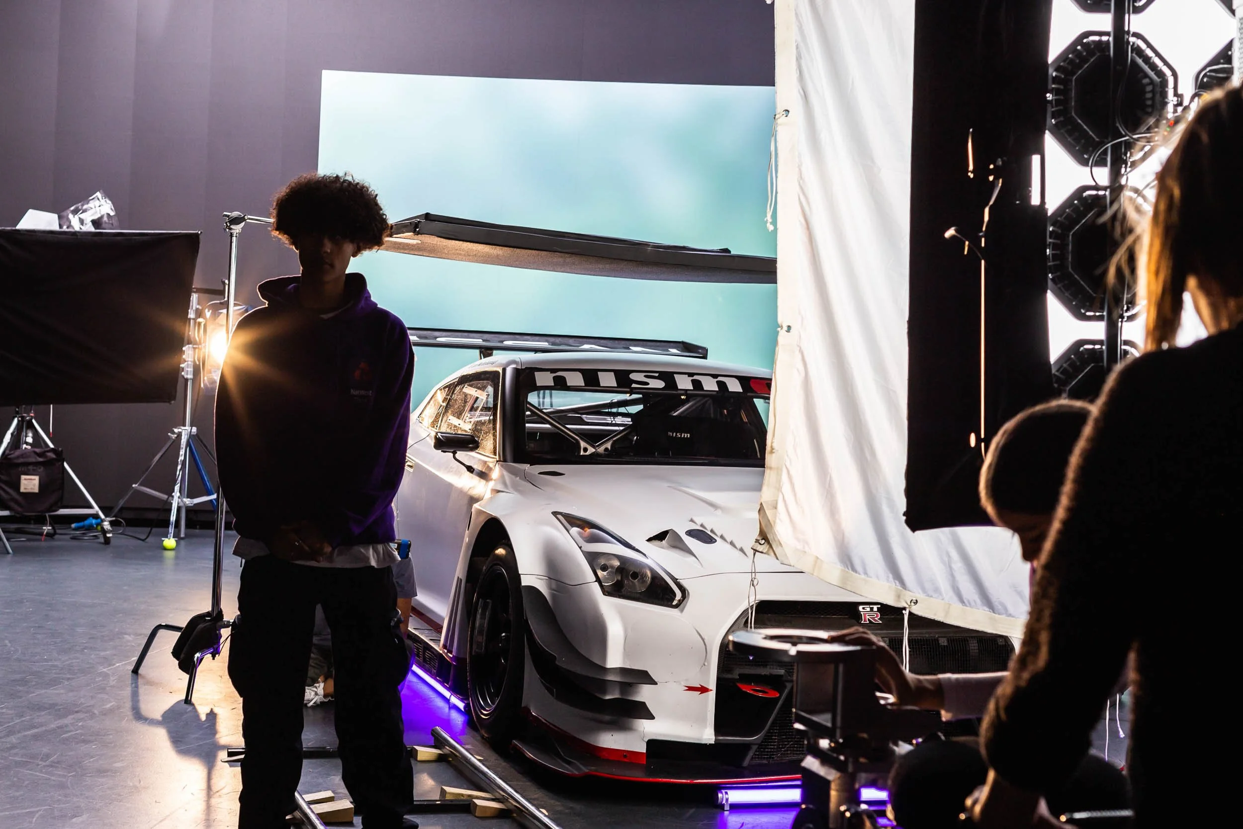 Production stills on set a young man walks towards camera with lights gleaming and a race car in the background
