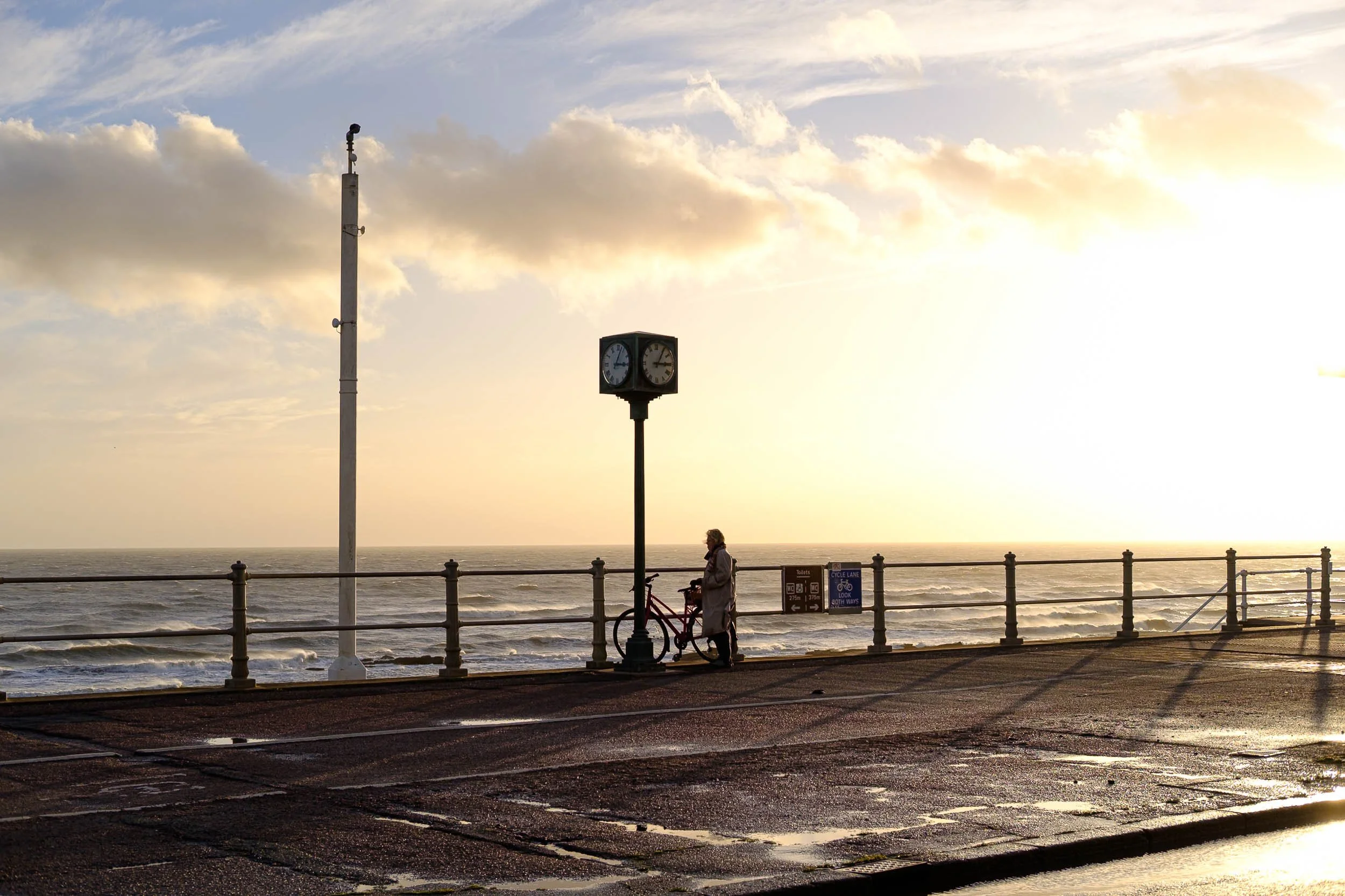 Golden hour at the beach on a windswept day a woman takes a rest with her bike by a giant street clock