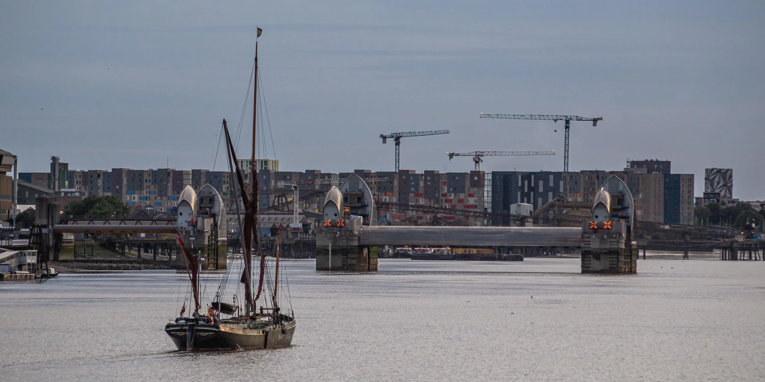 Thames Barrier in East London, E16. A sailboat approaches the barrier, with a view to Lewisham developments behind. Photography by Samuel Wheeler.