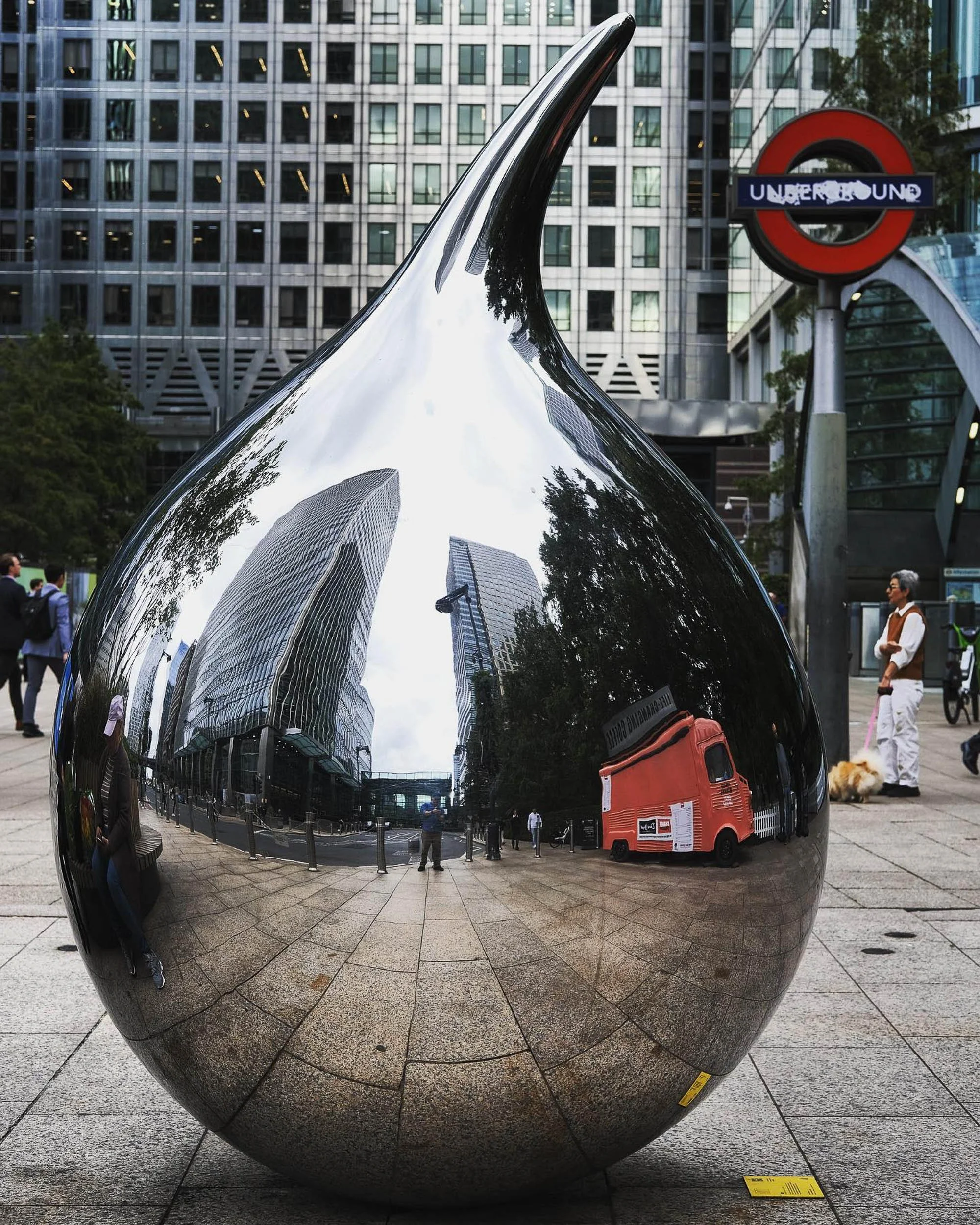 A shiny, reflective metallic sculpture located outdoors in an urban area, with a modern glass building and the London Underground sign in the background. People are walking nearby and the sculpture's surface reflects the surroundings.