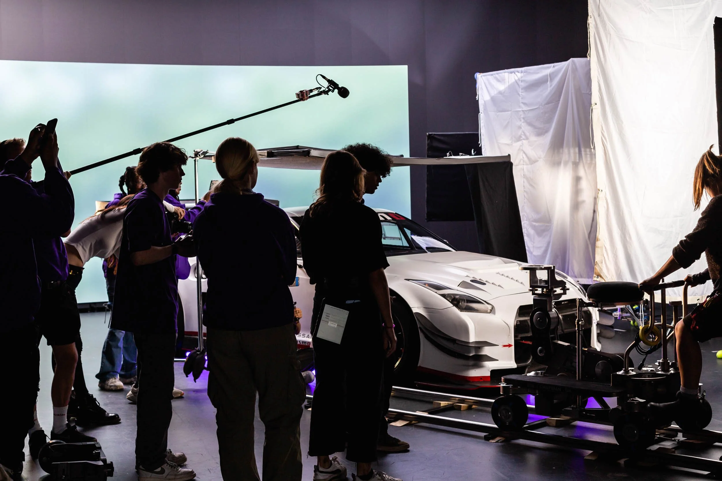 Group of people on set around a white sports car, with equipment including a camera crane and large led wall backdrop.