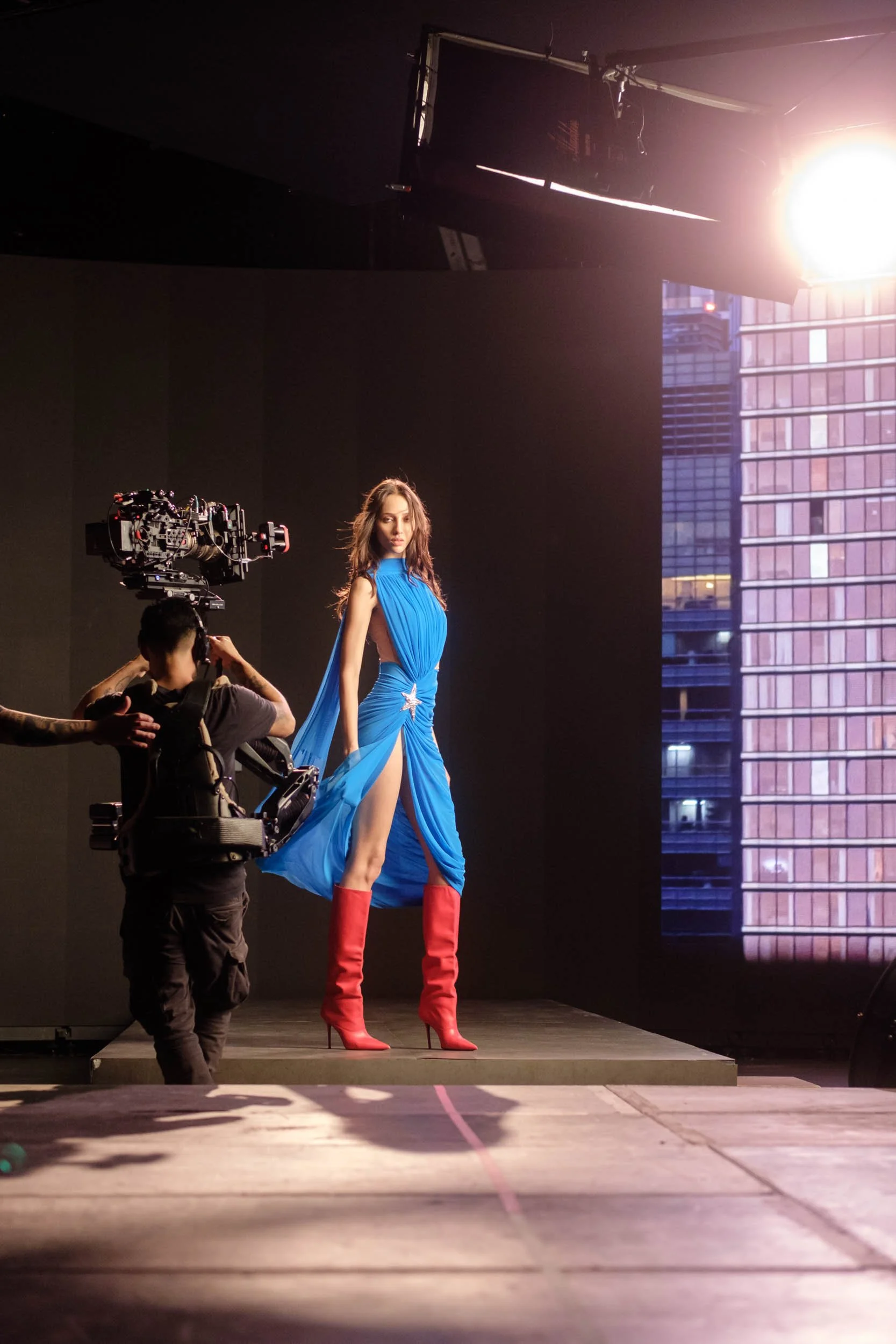 A woman wearing a blue gown and red high-heeled boots standing on a stage, being filmed by a crew member using a professional camera. The stage is indoors with a city skyline visible with large windows displayed on a large LED Screen.