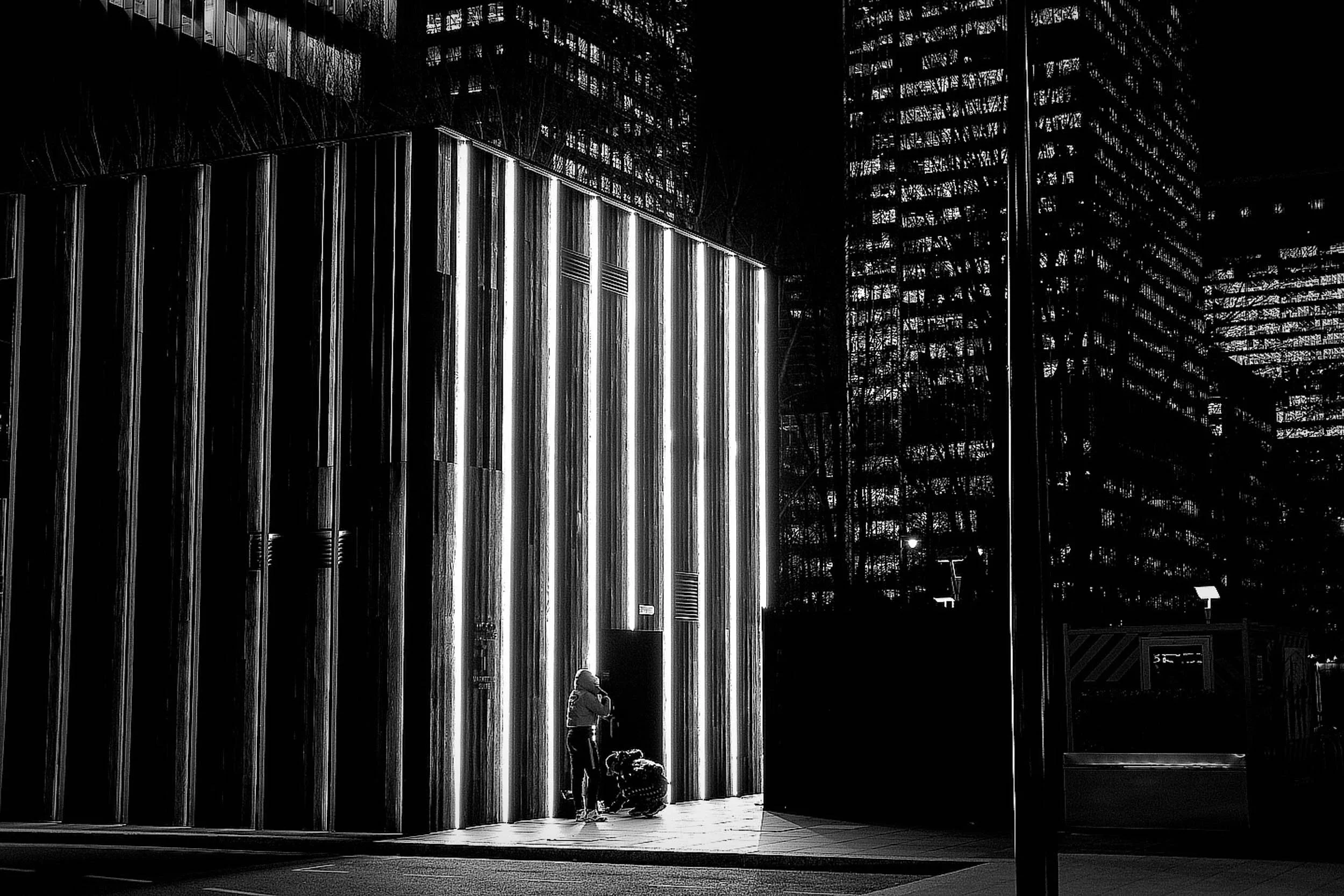 Black and white photo of two people at night in an urban setting, standing under large illuminated vertical metallic structures. Tall buildings with illuminated windows are visible in the background adding depth to the layers.