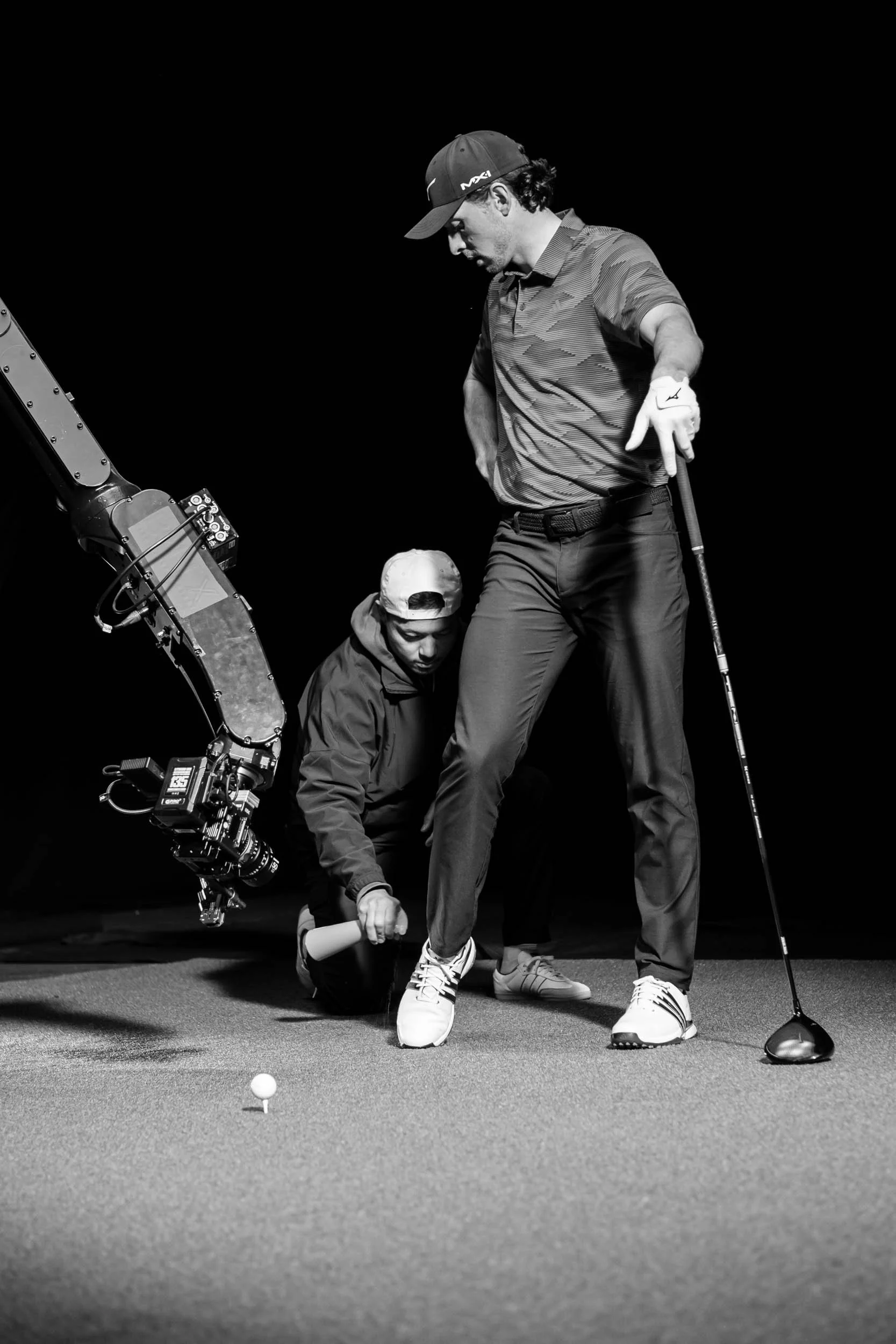striking high contrast black and white image of a male pro golfer lines up a shot as a production assistant prepares for next take with automated robotic camera arm